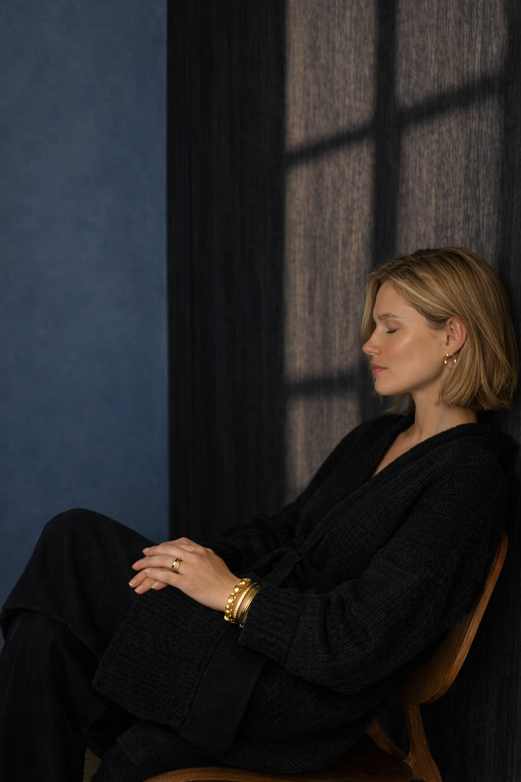 A woman with blonde hair sitting with her eyes closed against a dark textured wall, with sunlight casting shadows through window panes.