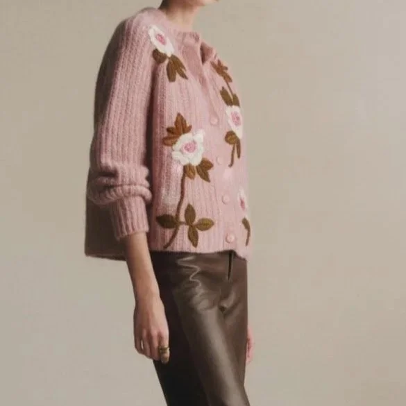 A woman wearing a pink knit cardigan with floral embroidery and dark pants standing against a plain wall.