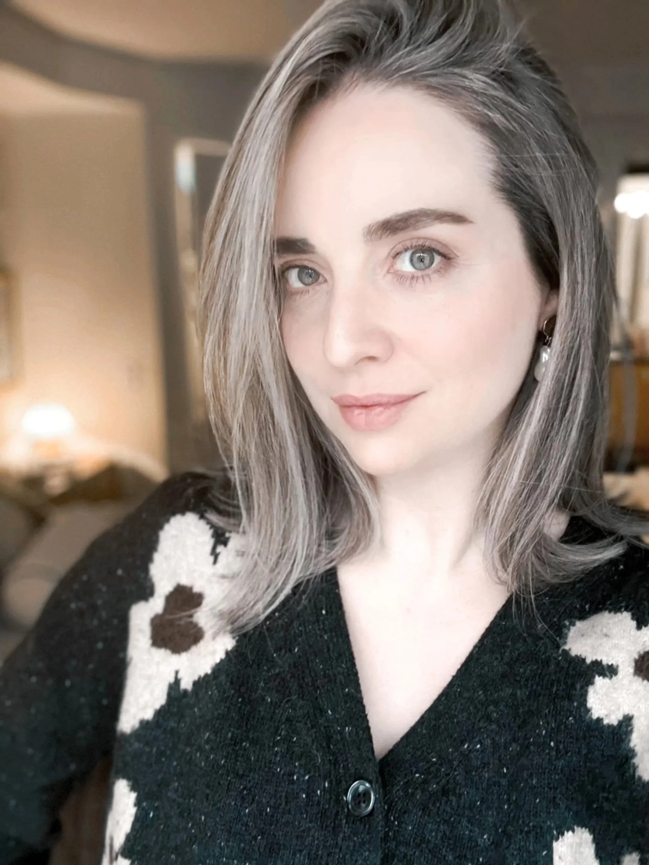 A woman with shoulder-length gray hair and blue eyes taking a selfie indoors. She is wearing a black sweater with white floral patterns and pearl earrings.