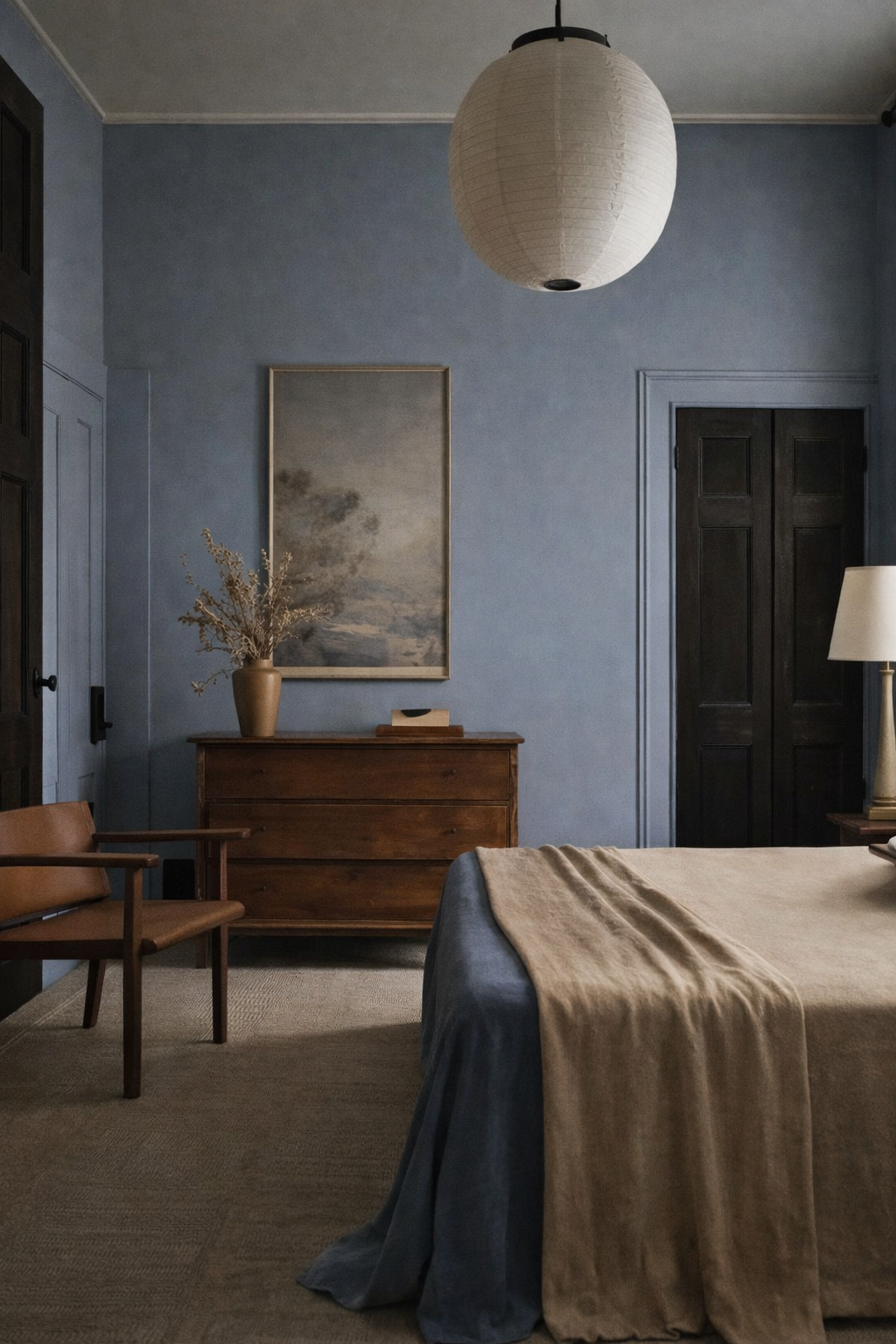 A cozy bedroom with muted blue walls, a wooden dresser with a beige vase and dried flowers, a framed landscape painting, a tan chair, a bed with a beige blanket, a lamp, and a paper lantern ceiling light.