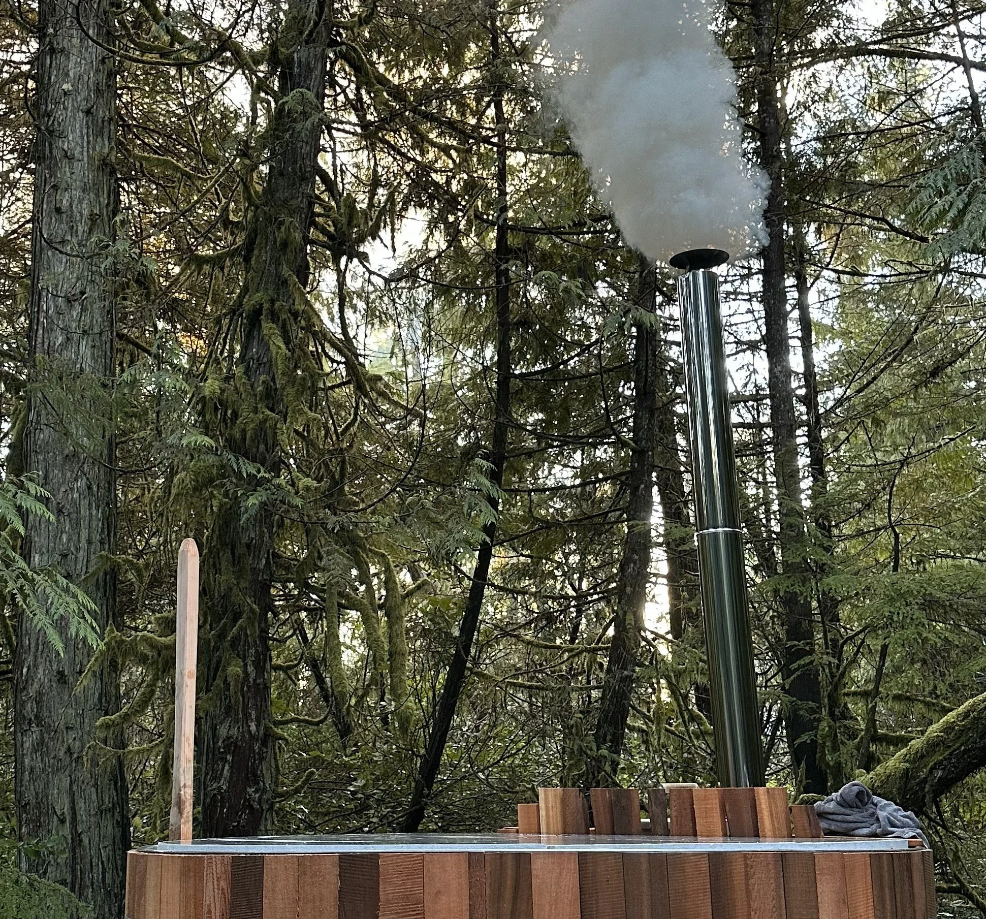 Wood fired hot tub in forest setting with light filtering through trees and smoke pouring out of shiny chimney