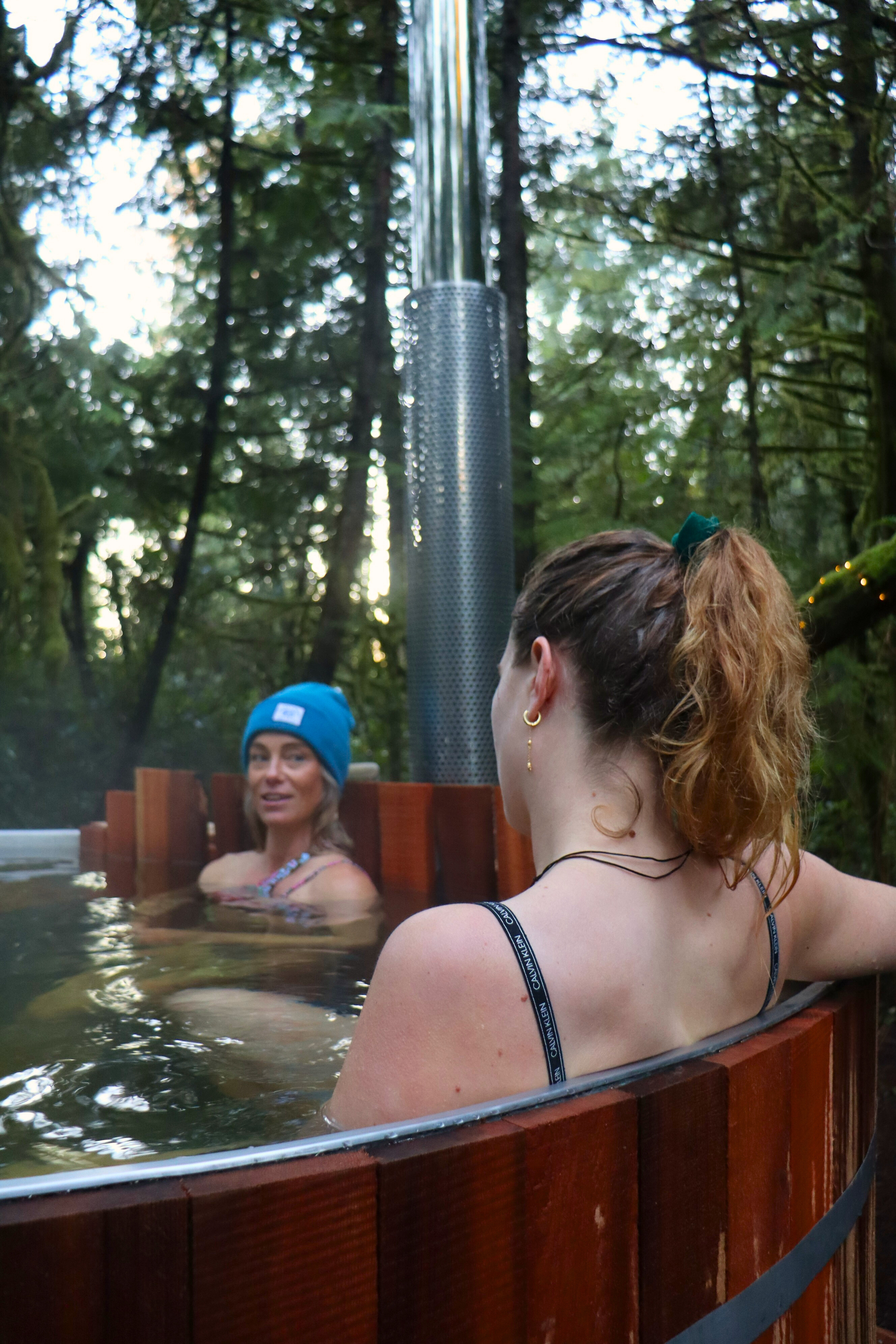 two women sitting in woodfired hot tub with shiny chimney and forest in the background with sunset light
