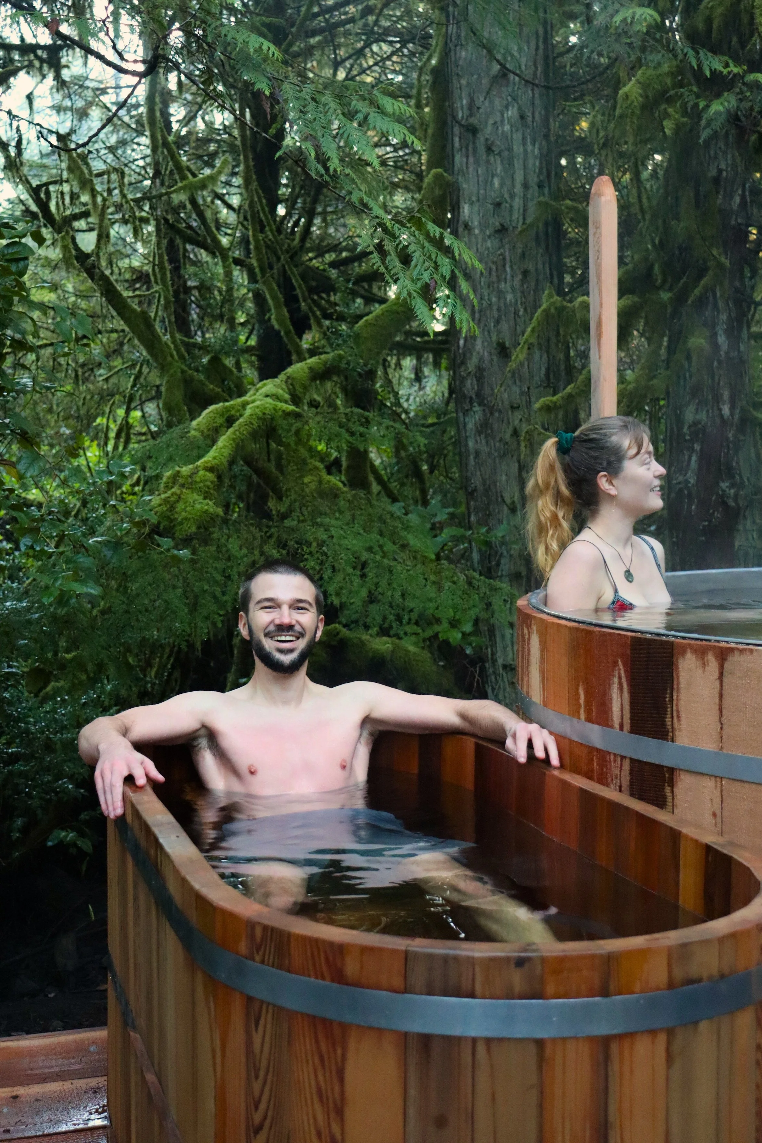 Tofino hot tub and cold plunge with man sitting in cold plunge smiling, woman in hot tub smiling, with green rainforest and moss
