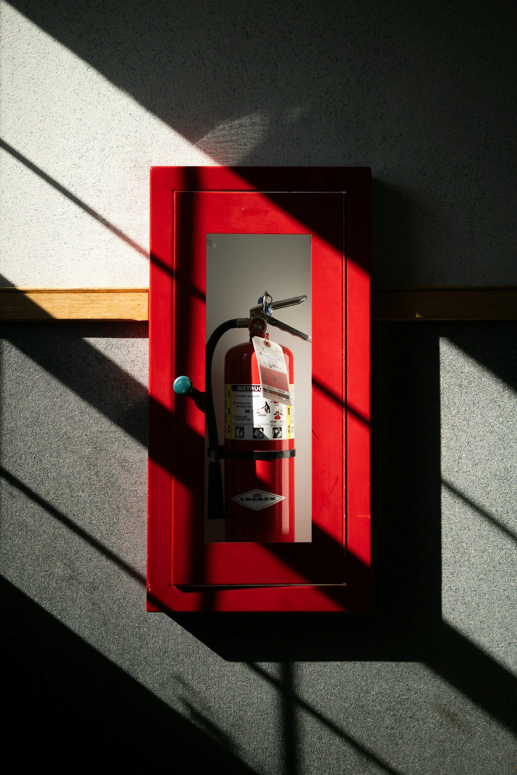 Red fire alarm pull station with a fire extinguisher inside a glass case, mounted on a wall with light and shadow patterns.