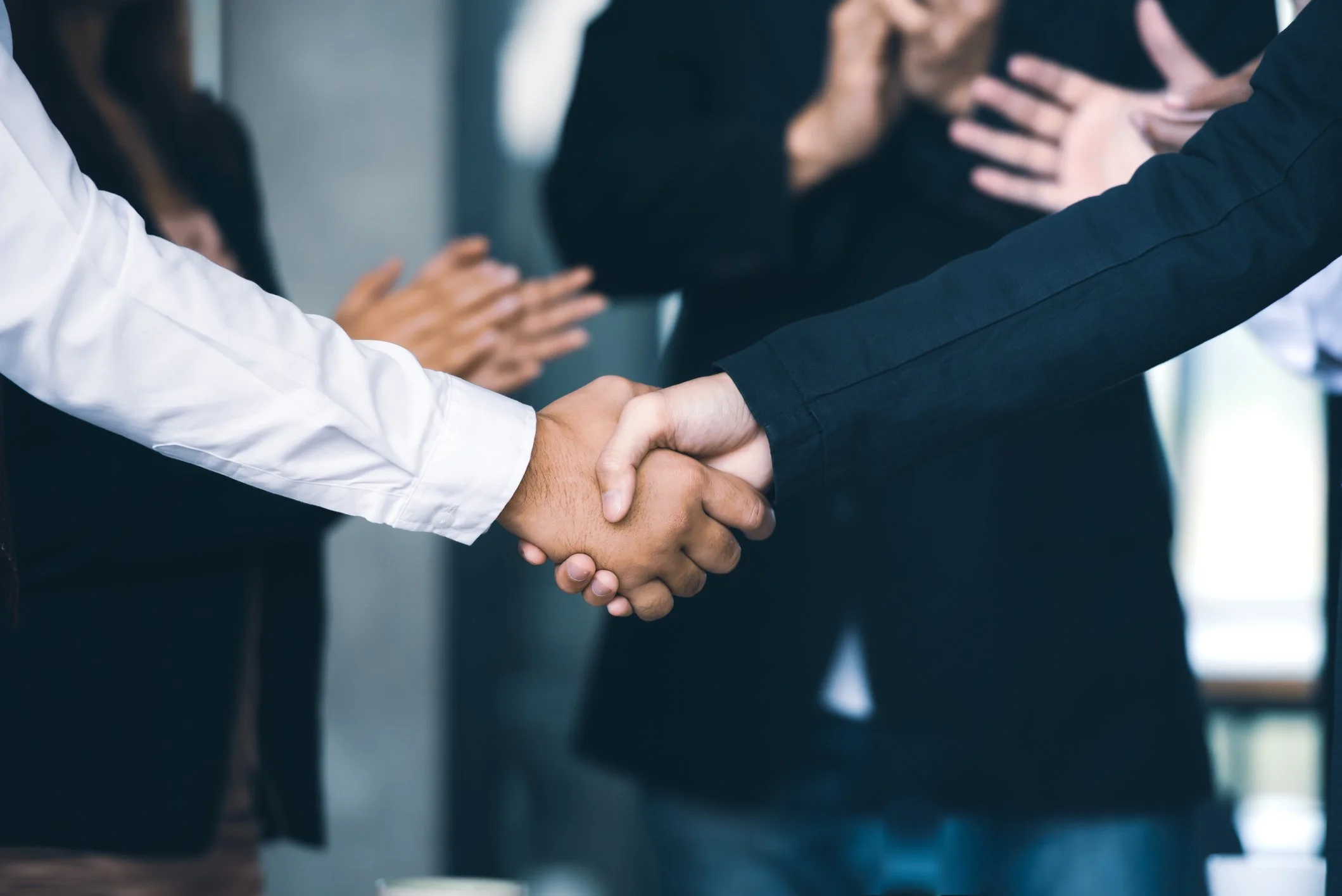 Two people shaking hands with a group clapping in the background, in a professional setting.