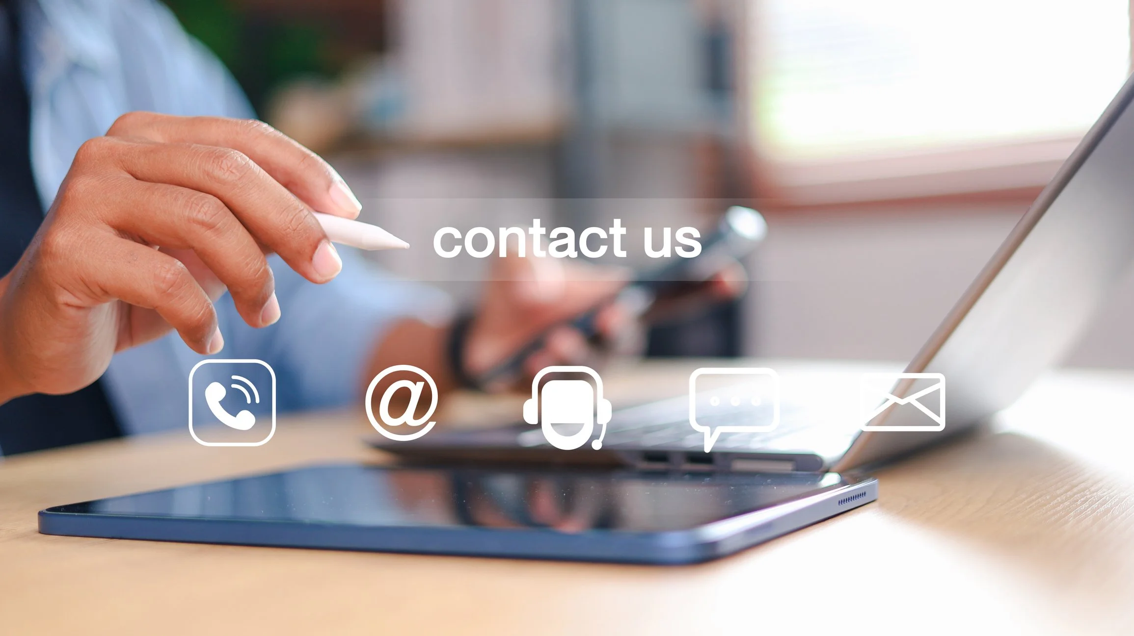 Person holding a stylus and using a smartphone with contact icons for phone, email, headset, chat, and mail overlaid on the image, alongside a tablet on a wooden desk.