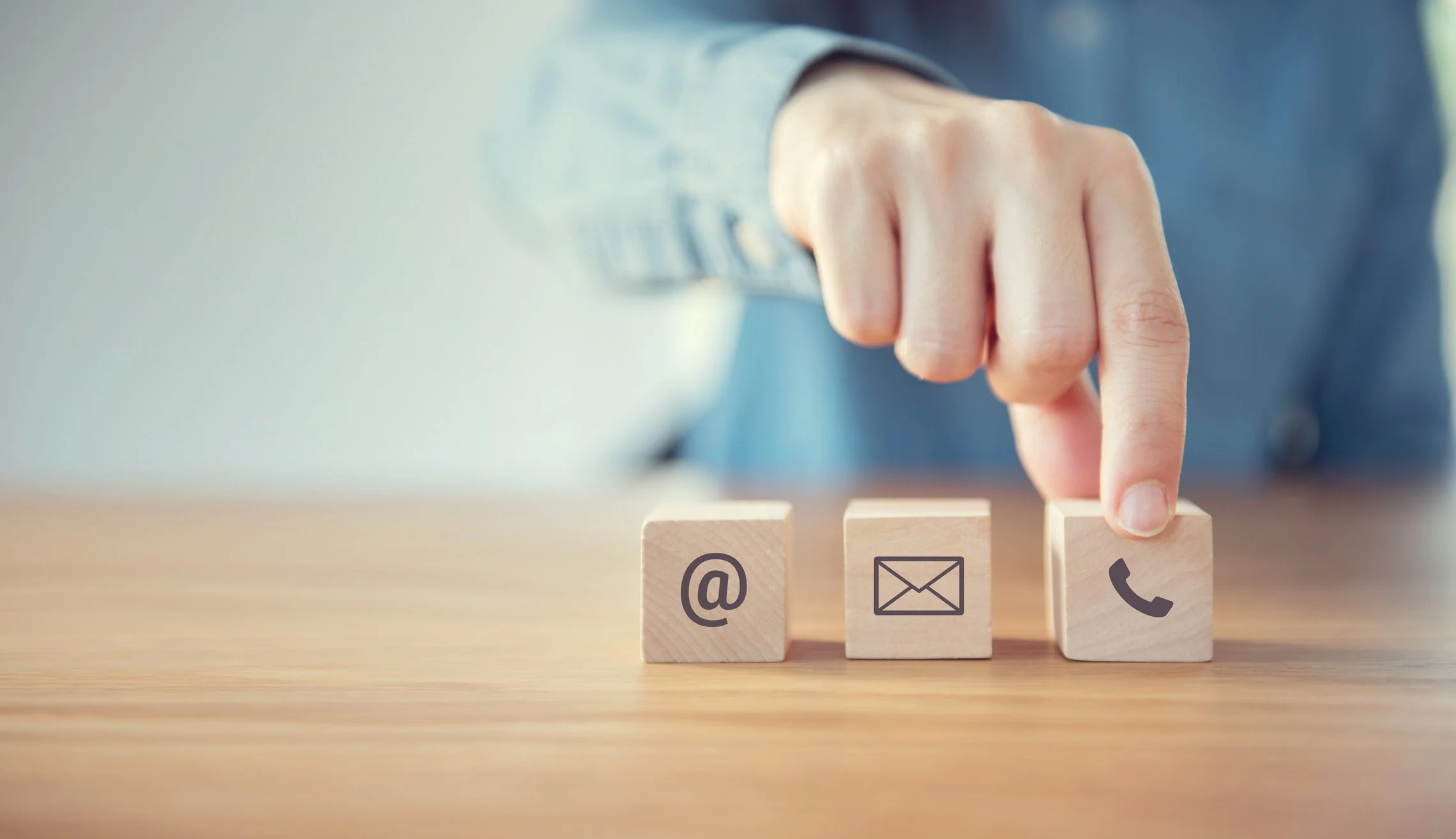 A person's hand pressing a wooden block with a phone icon, with additional wooden blocks showing email and at symbols on a wooden table.