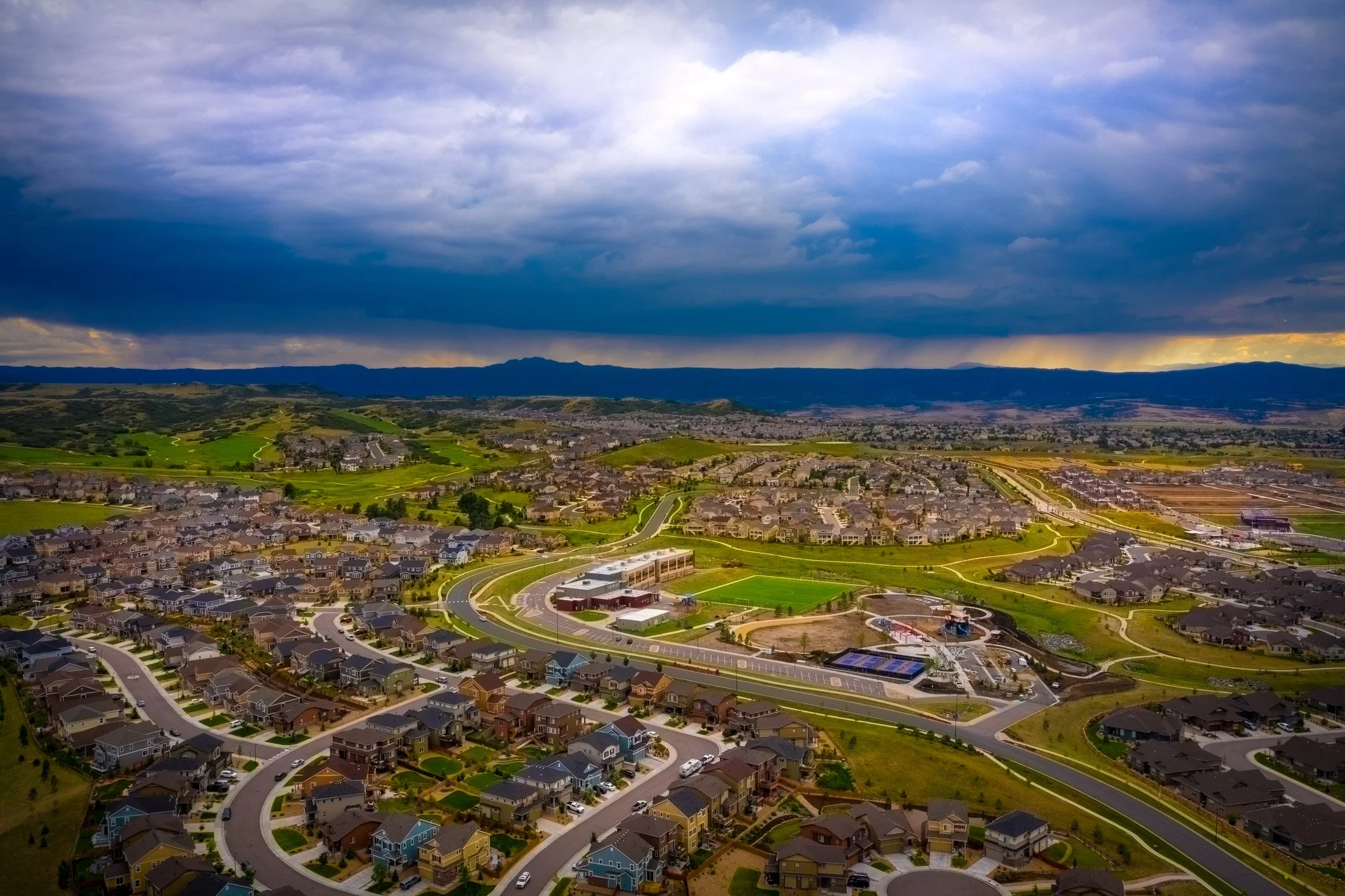 Aerial view of a suburban neighborhood with winding streets, houses, a park, and a sports field under a cloudy sky.
