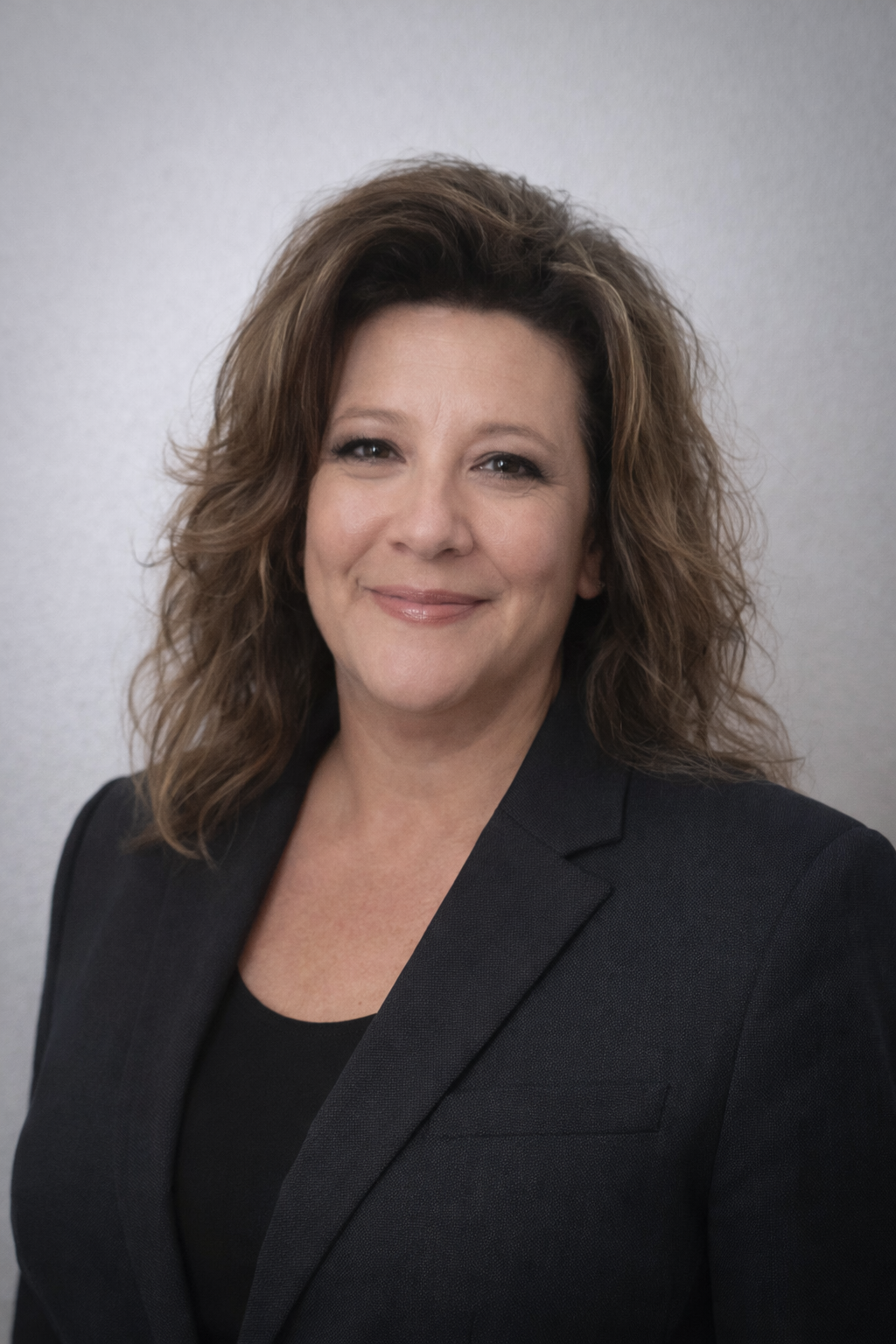 Julie Baca - A professional woman with wavy brown hair smiling, wearing a black blazer, standing against a plain gray wall.