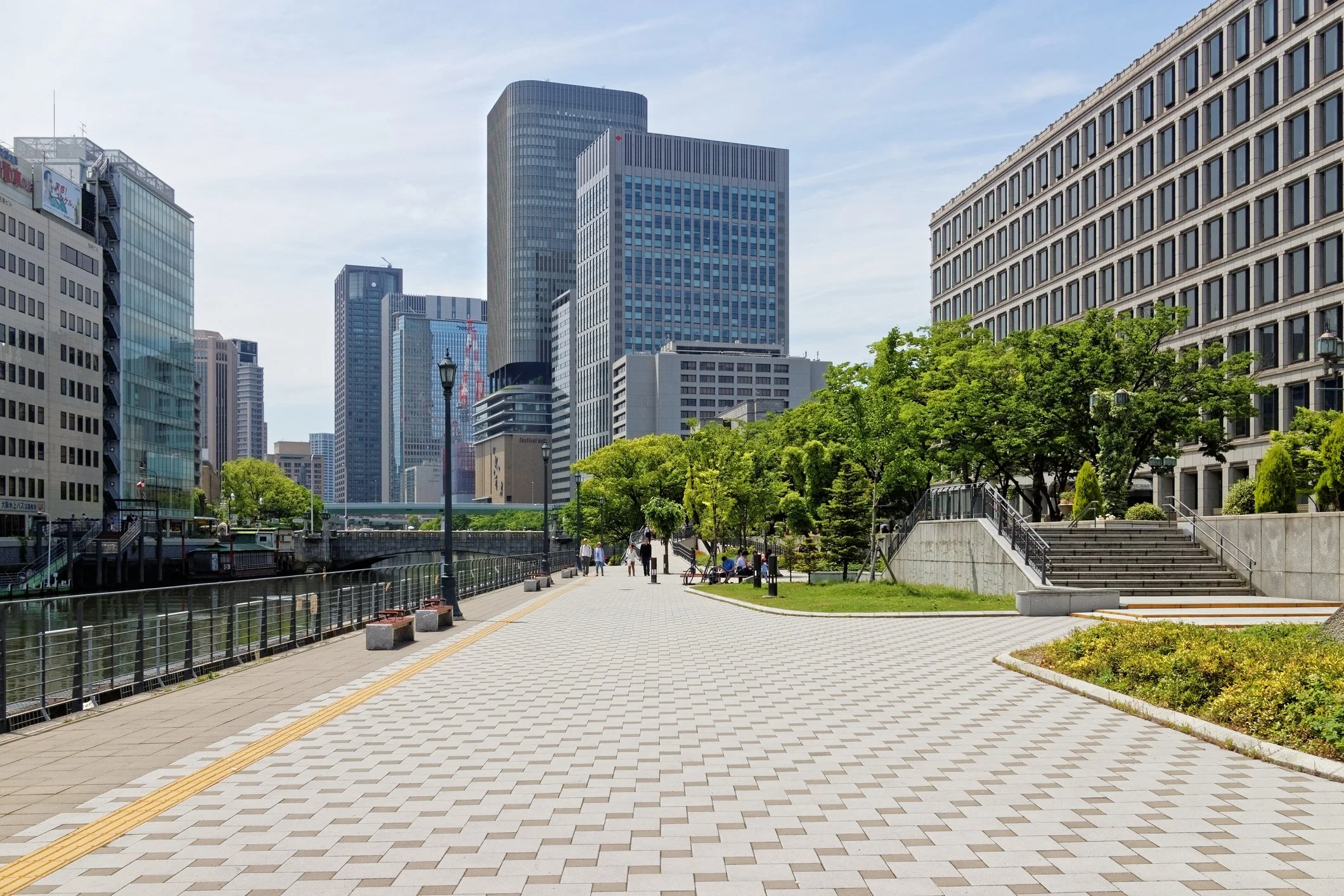 A cityscape with modern high-rise buildings and a riverside promenade with trees and benches.