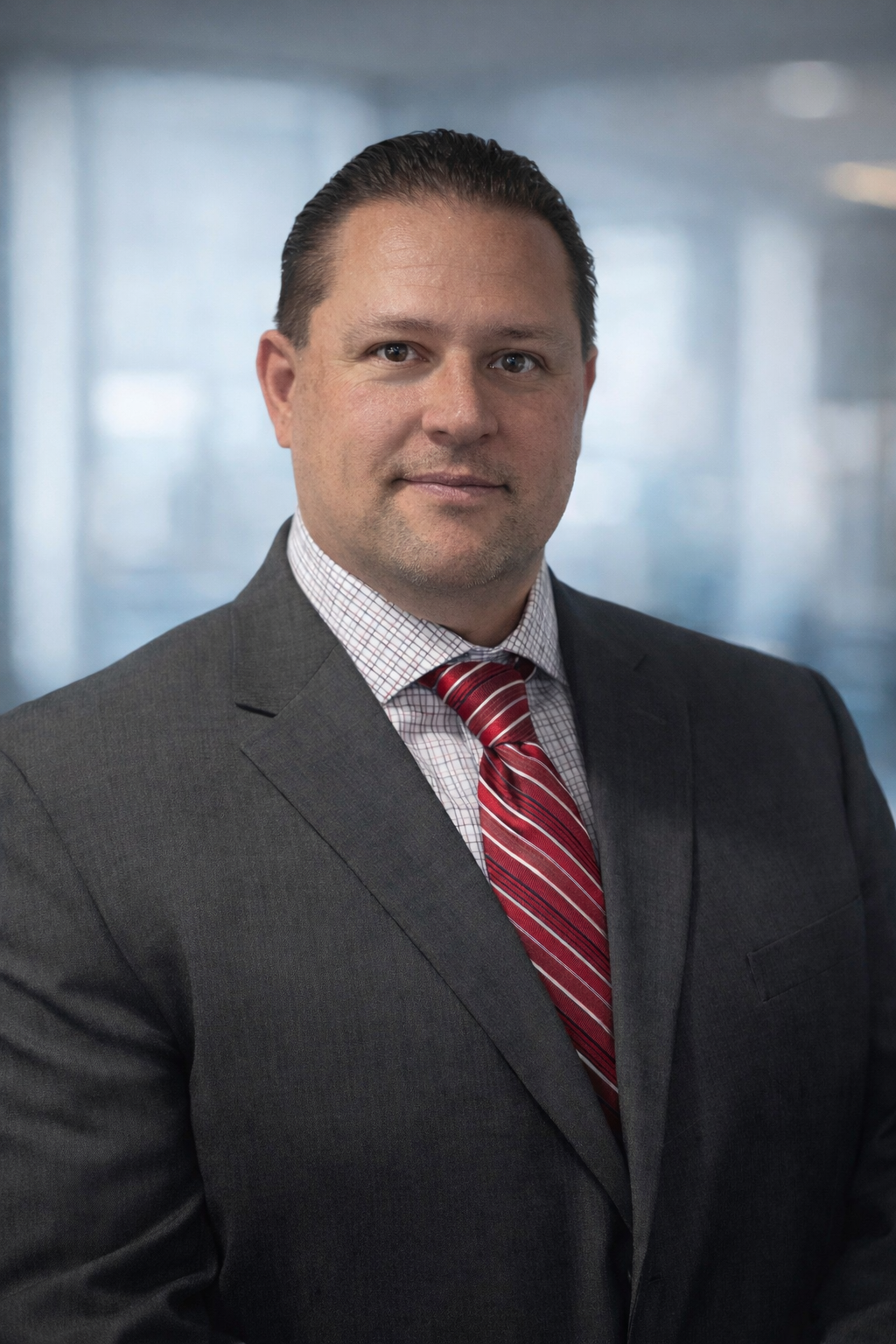 Adam Bryning, CMCA, AMS - A professional man wearing a gray suit, a red striped tie, and a checkered shirt posing in an office setting with a blurred background.