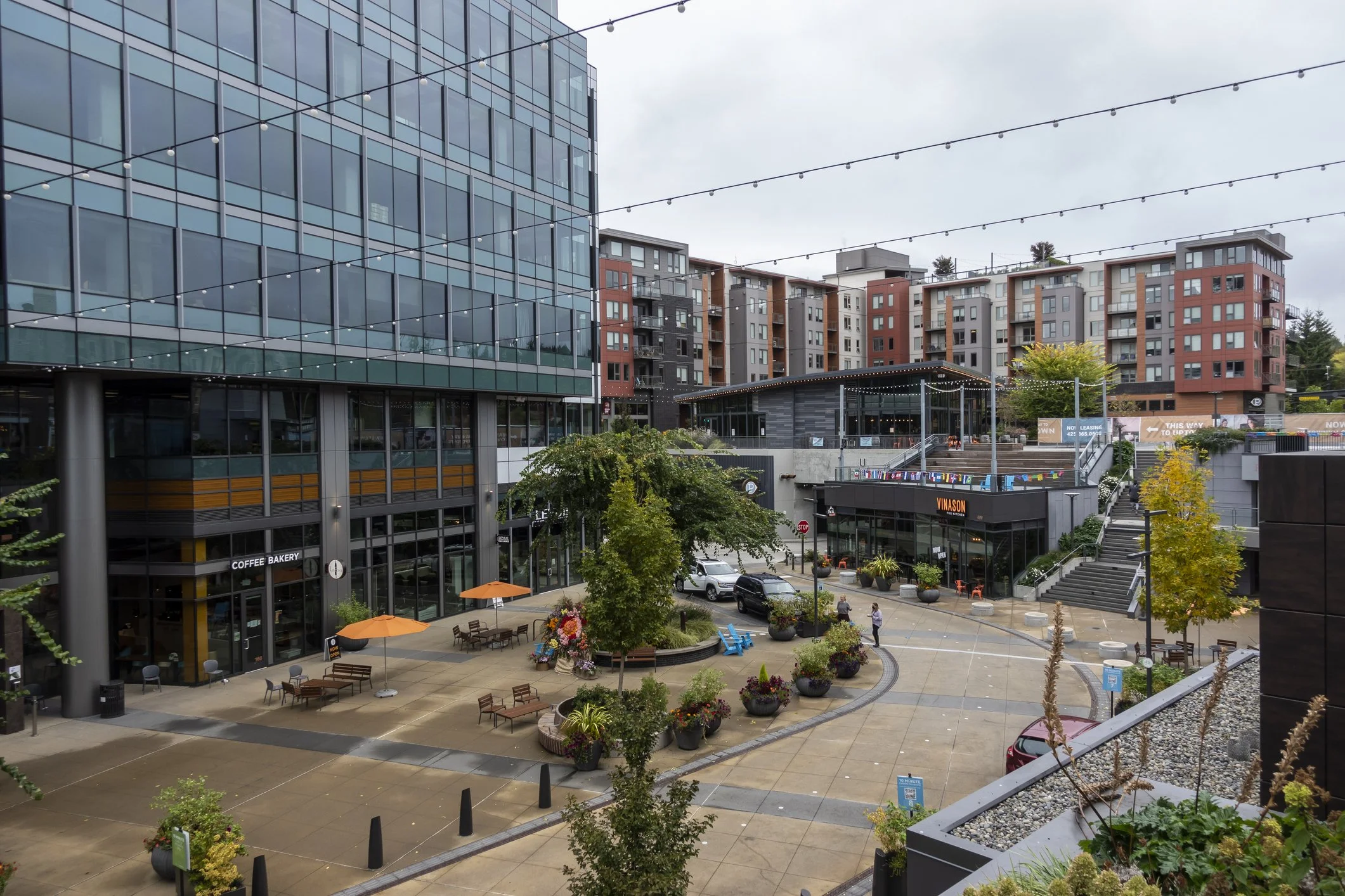 A modern urban plaza with outdoor seating, potted plants, trees, and a coffee shop, surrounded by multi-story residential and commercial buildings during daytime with overcast sky.