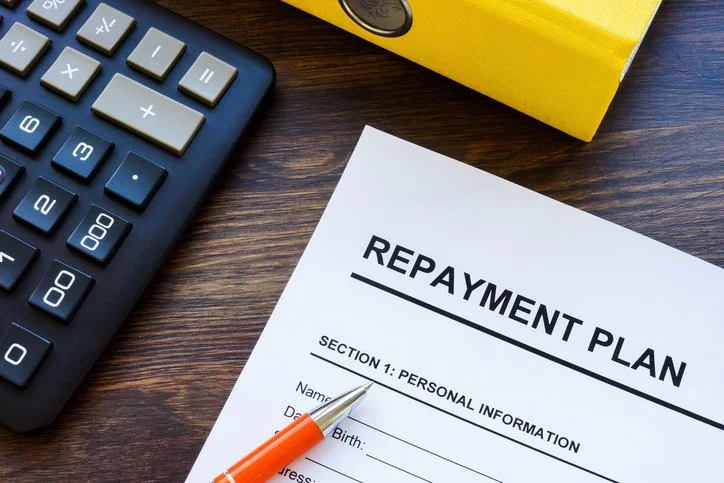 A repayment plan document on a wooden desk with a pen, calculator, and yellow box nearby.