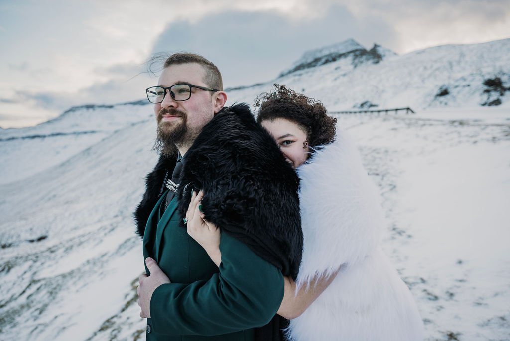 A couple standing close together outdoors in a snowy mountainous landscape, with the woman hugging the man from behind and partially hidden behind his shoulder.