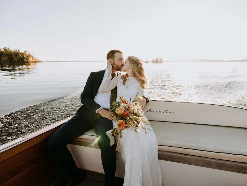 A bride and groom sharing a kiss on a boat, with water and a distant shoreline in the background, during sunset at Migis Lodge, Maine