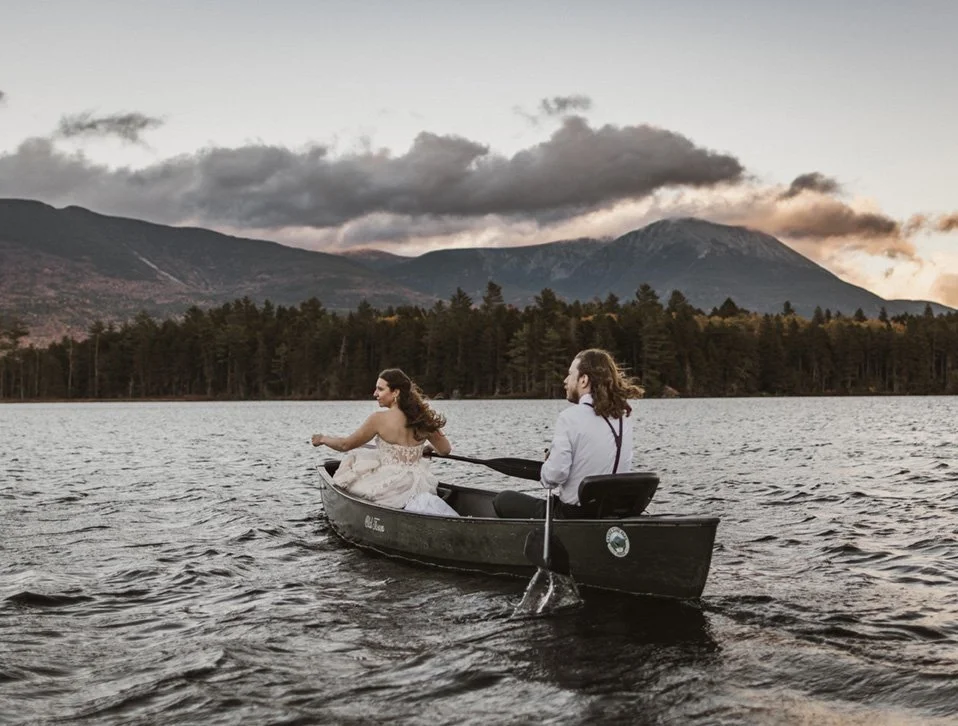 A man and woman holding hands and smiling at each other in an open grassy field with mountains in the background.