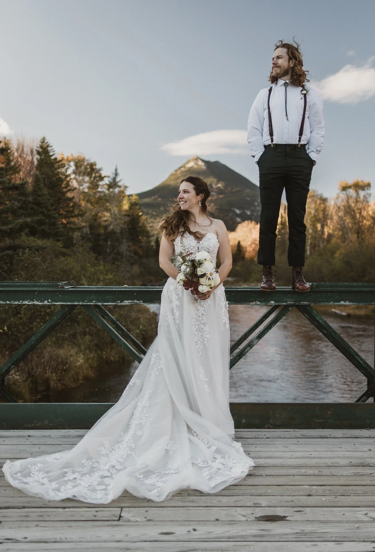 A bride in a wedding dress holding a bouquet near a river, with a groom standing on a bridge above her, mountains and trees in the background during sunset.