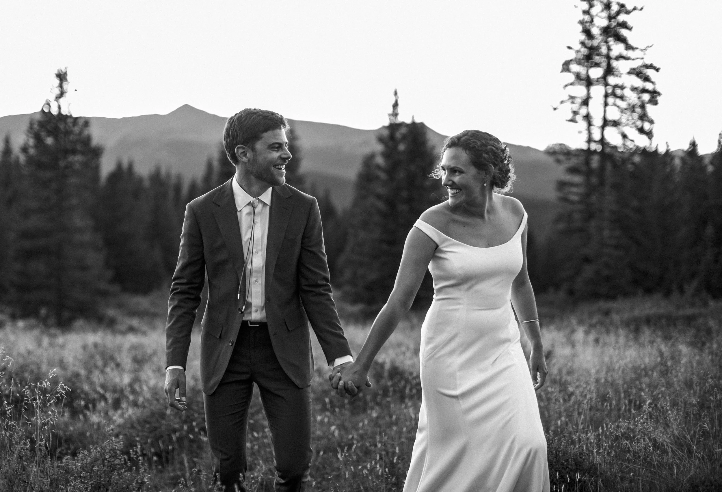 A couple in wedding attire walking hand in hand outdoors in a field with trees and mountains in the background in Leadville, Colorado, smiling at each other, black and white photo.