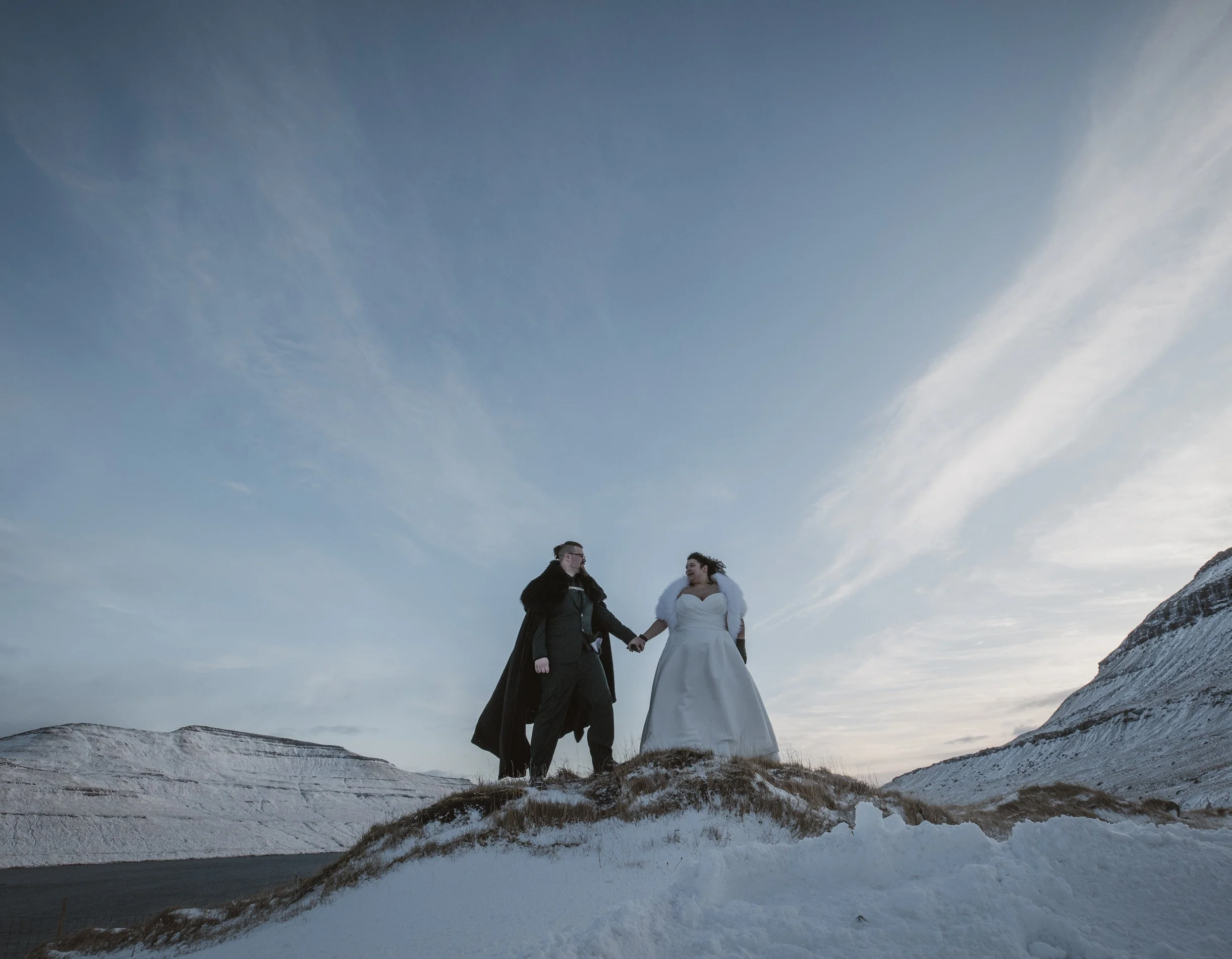 A couple dressed in wedding attire holding hands on a snowy hilltop with mountains and sky in the background at sunset in the Faroe Islands