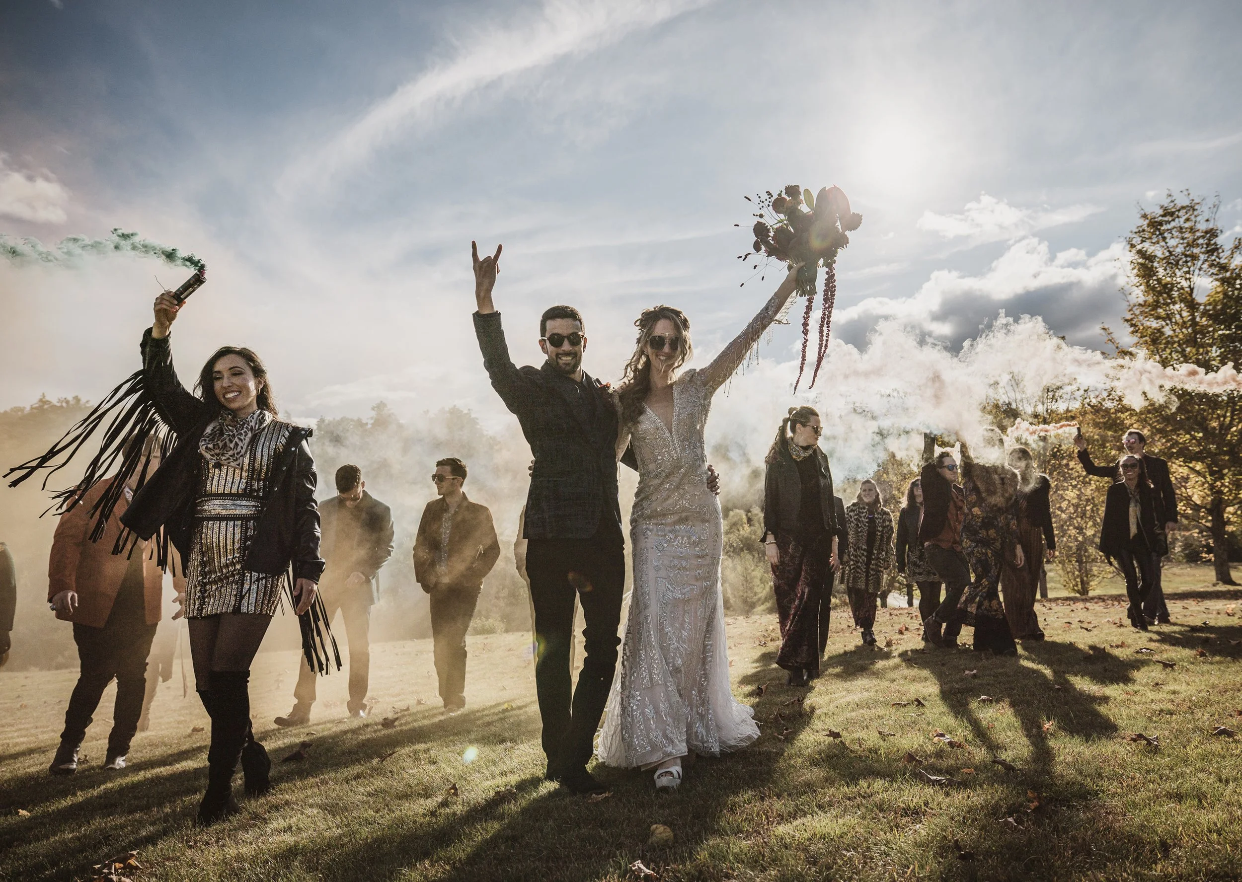 A bride and groom celebrating outdoors with friends, holding smoke flares and gift, in a park on a sunny day.