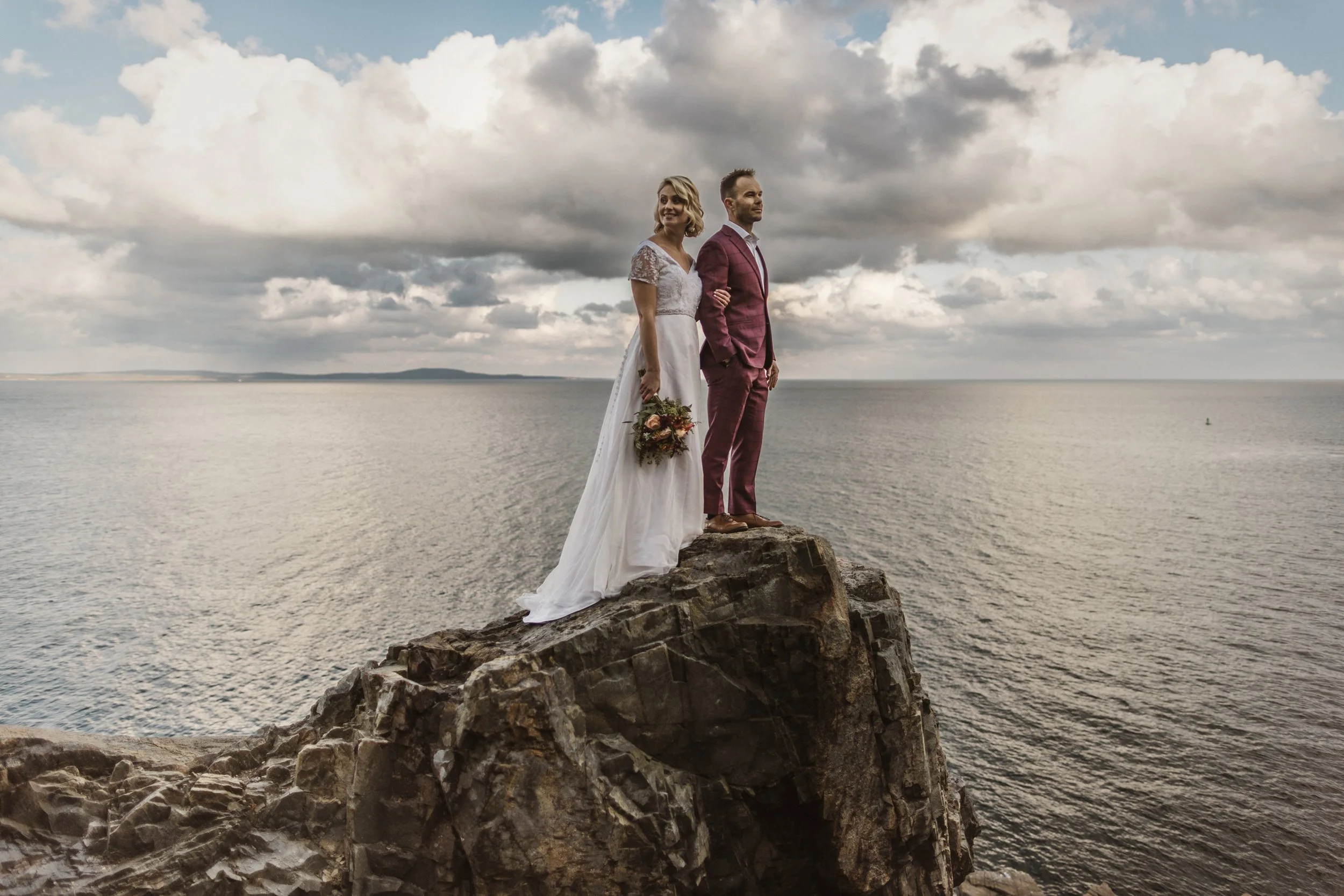 A bride and groom stand on a rocky outcrop in Acadia National Park overlooking the ocean on a cloudy day, with the bride holding a bouquet of flowers.