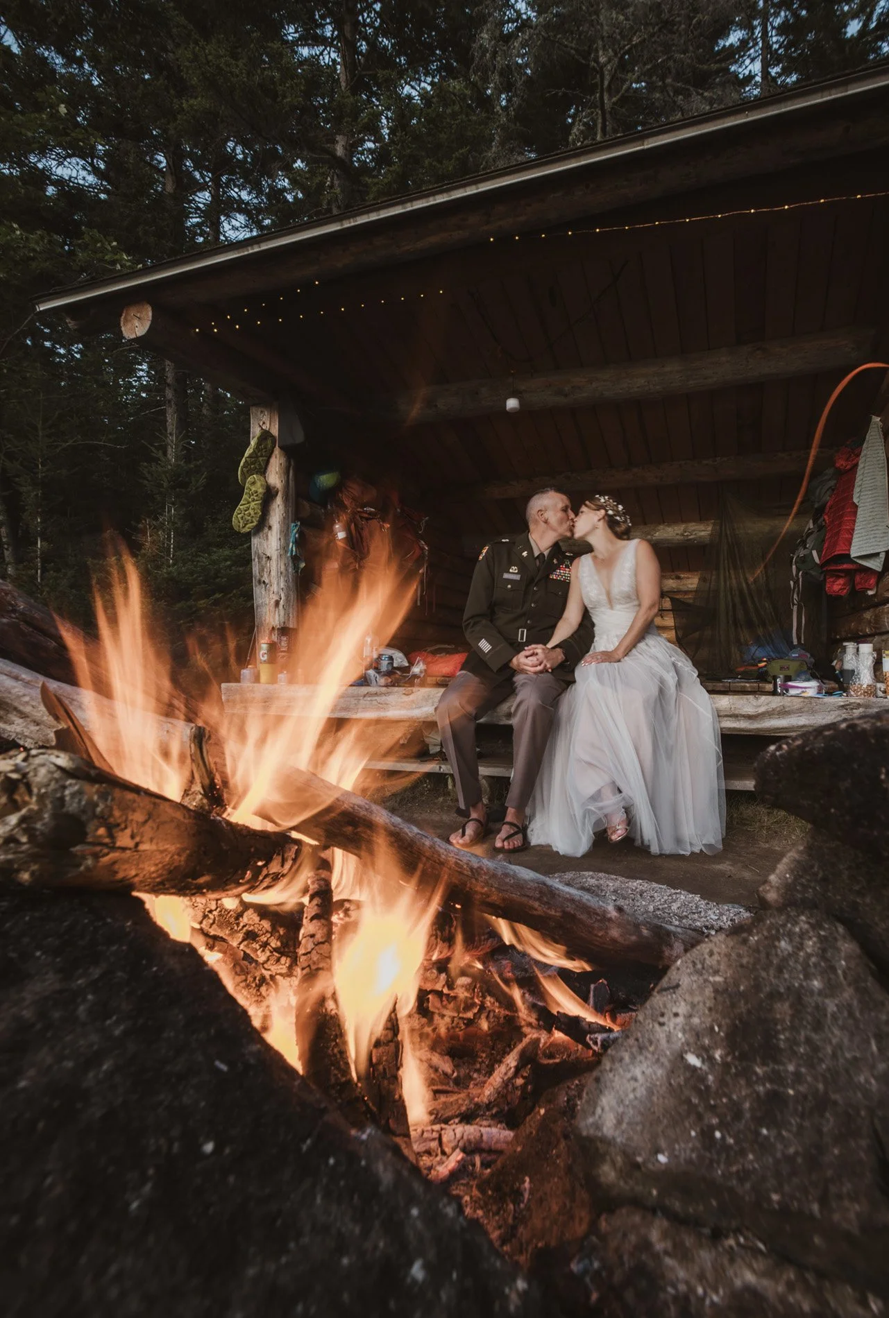 A couple sitting on a wooden platform inside a rustic shelter in the woods in Baxter State Park, Maine, kissing, with a campfire burning in the foreground and trees surrounding the area.