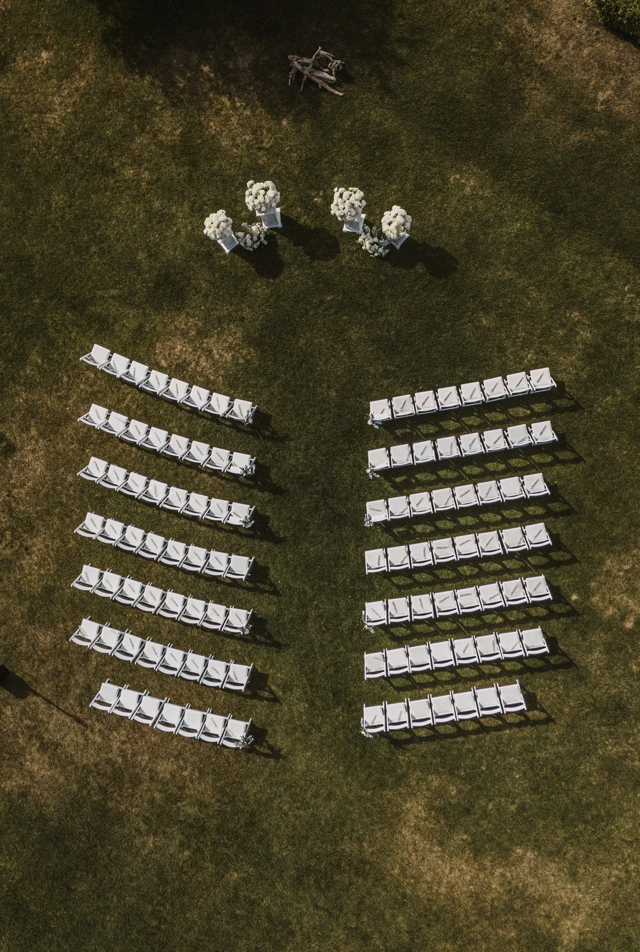 An aerial view of a wedding setup on grass, featuring rows of white chairs, floral arrangements, and a wooden bench.