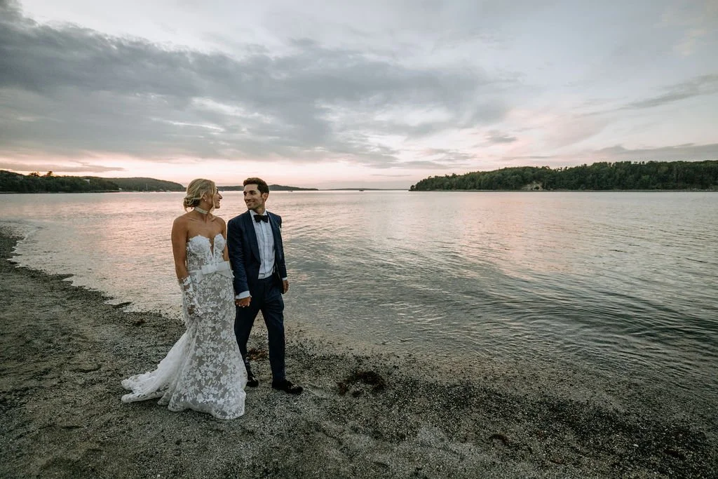 A bride in a white lace wedding dress and groom in a navy suit with a bow tie holding hands on a beach by a lake during sunset.