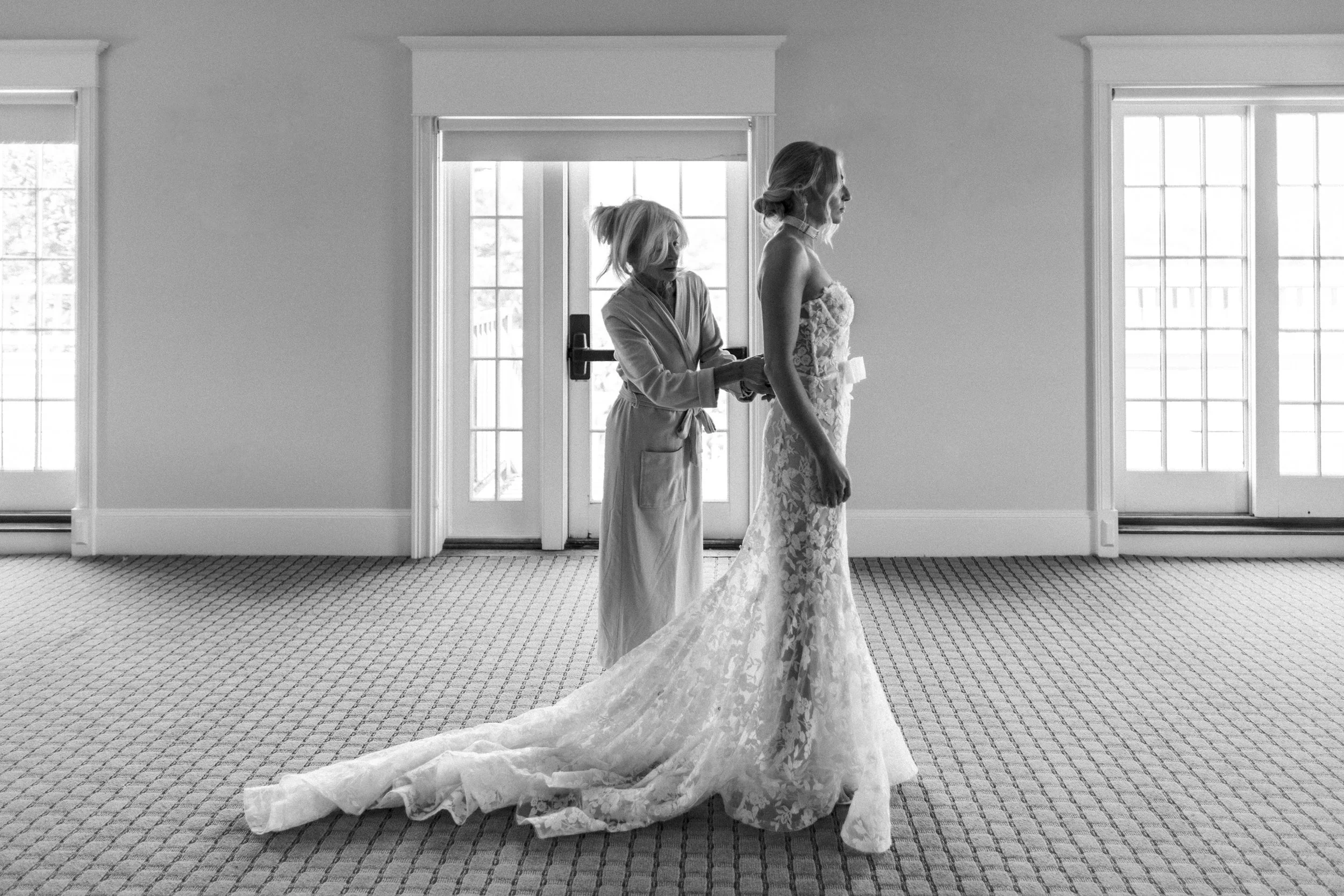 A woman in a wedding dress stands as another woman helps her zip or fasten the dress in a room with large windows and a patterned carpet.