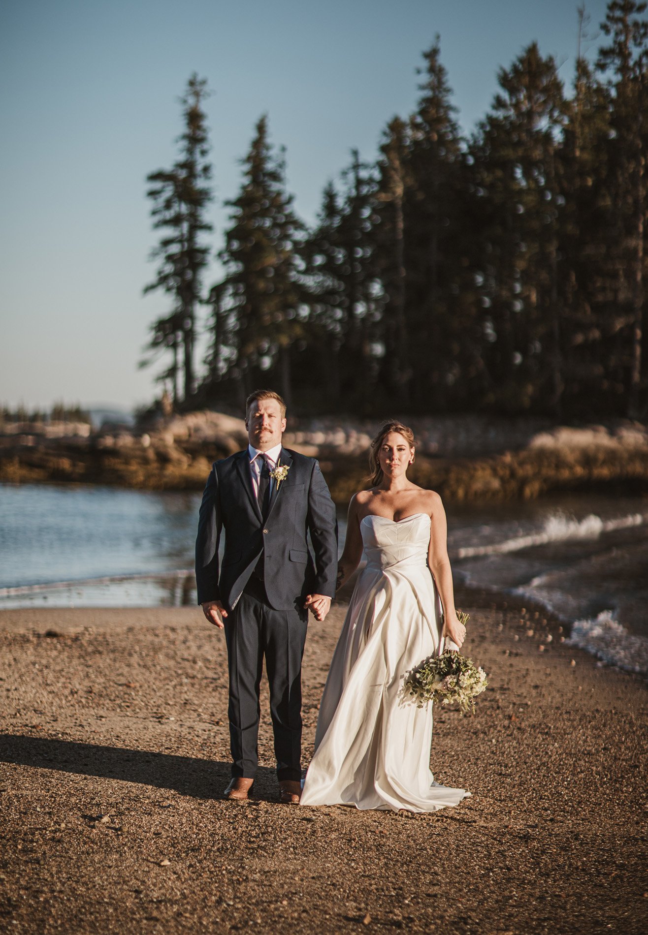 A bride and groom walking hand in hand on a sandy beach in Acadia National Park during sunset. The bride is in a strapless white wedding dress holding a bouquet, and the groom is in a dark suit with a tie and boutonniere.