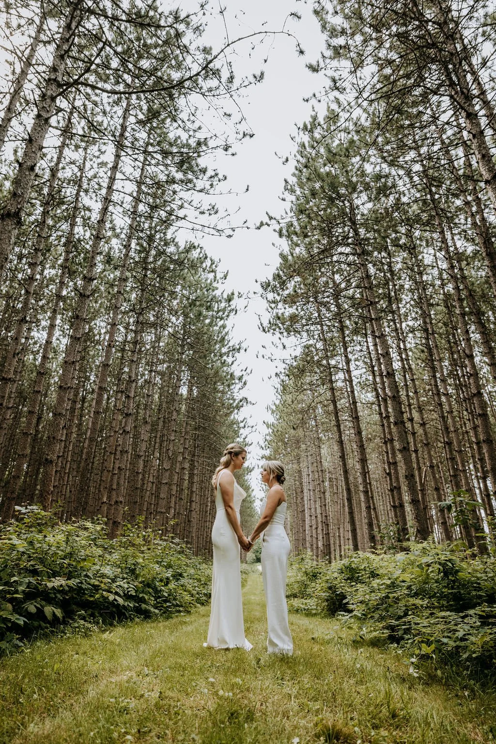 Two women in white dresses holding hands and facing each other in a forest with tall trees and green foliage at Cunningham Farm, Maine