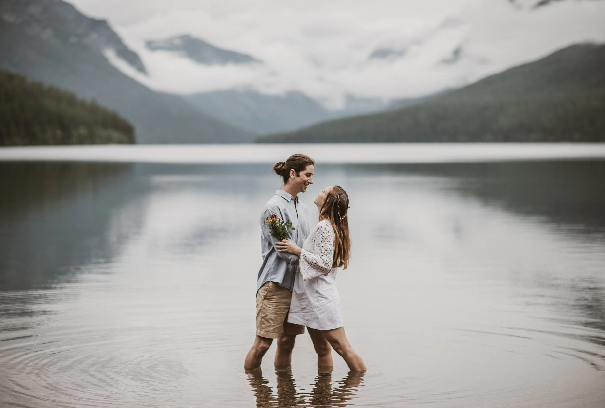A couple standing in a lake in Glacier National Park, Montana, exchanging a loving look, with a scenic mountain landscape in the background.