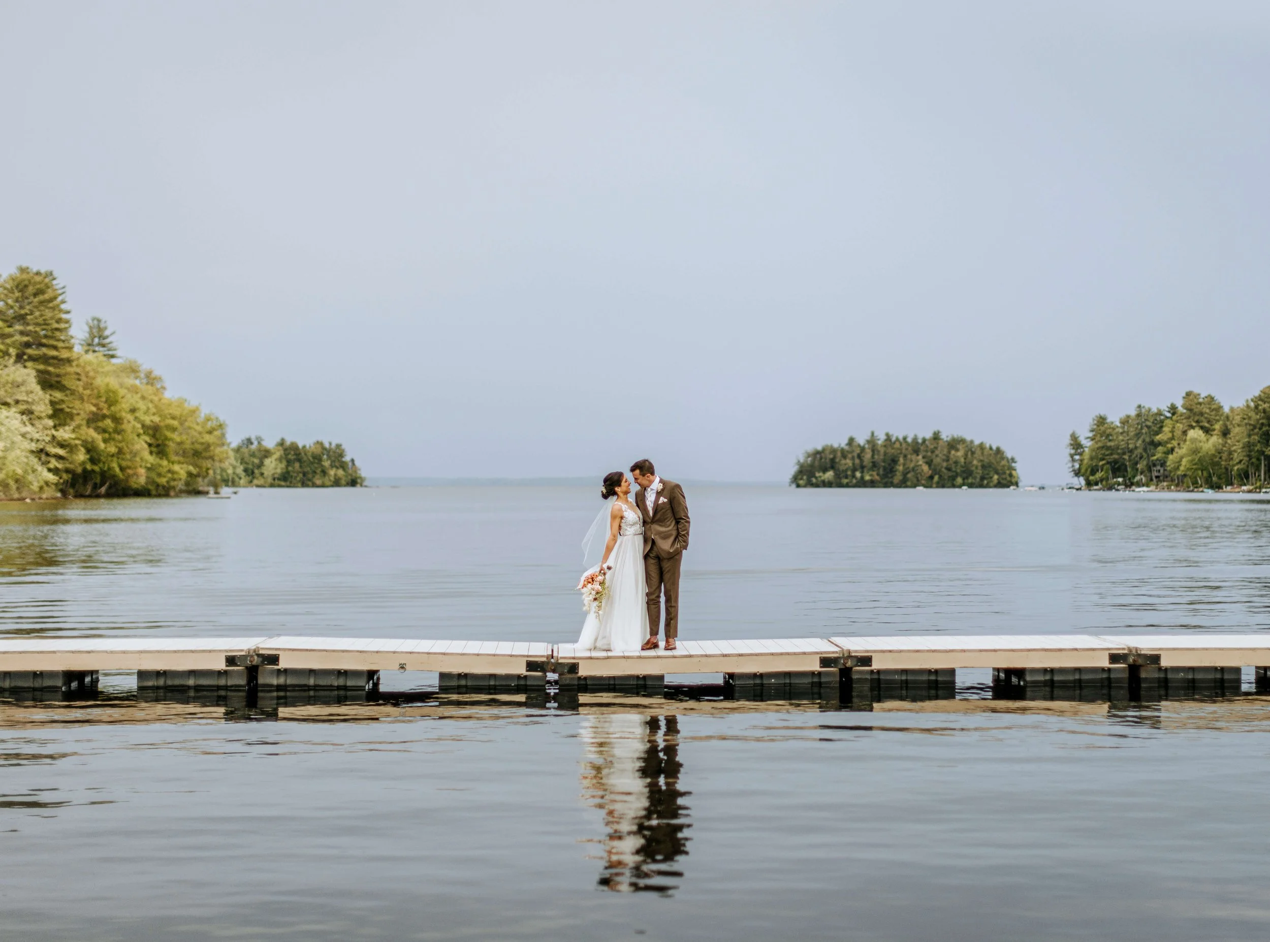 A bride and groom stand close together on a wooden dock by a calm lake, surrounded by trees, with a light overcast sky.
