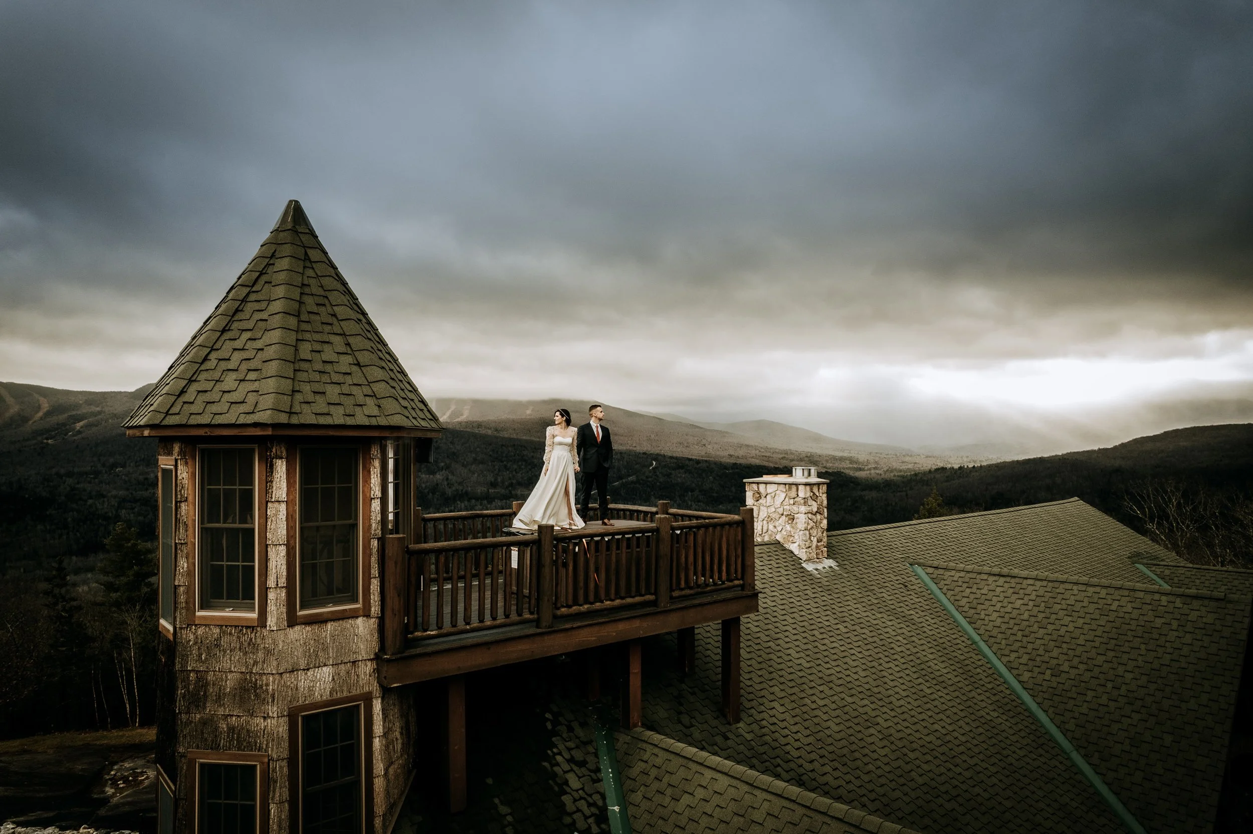 A bride and groom standing on a wooden balcony of a mountain house, with dark stormy clouds in the sky and mountains in the background.