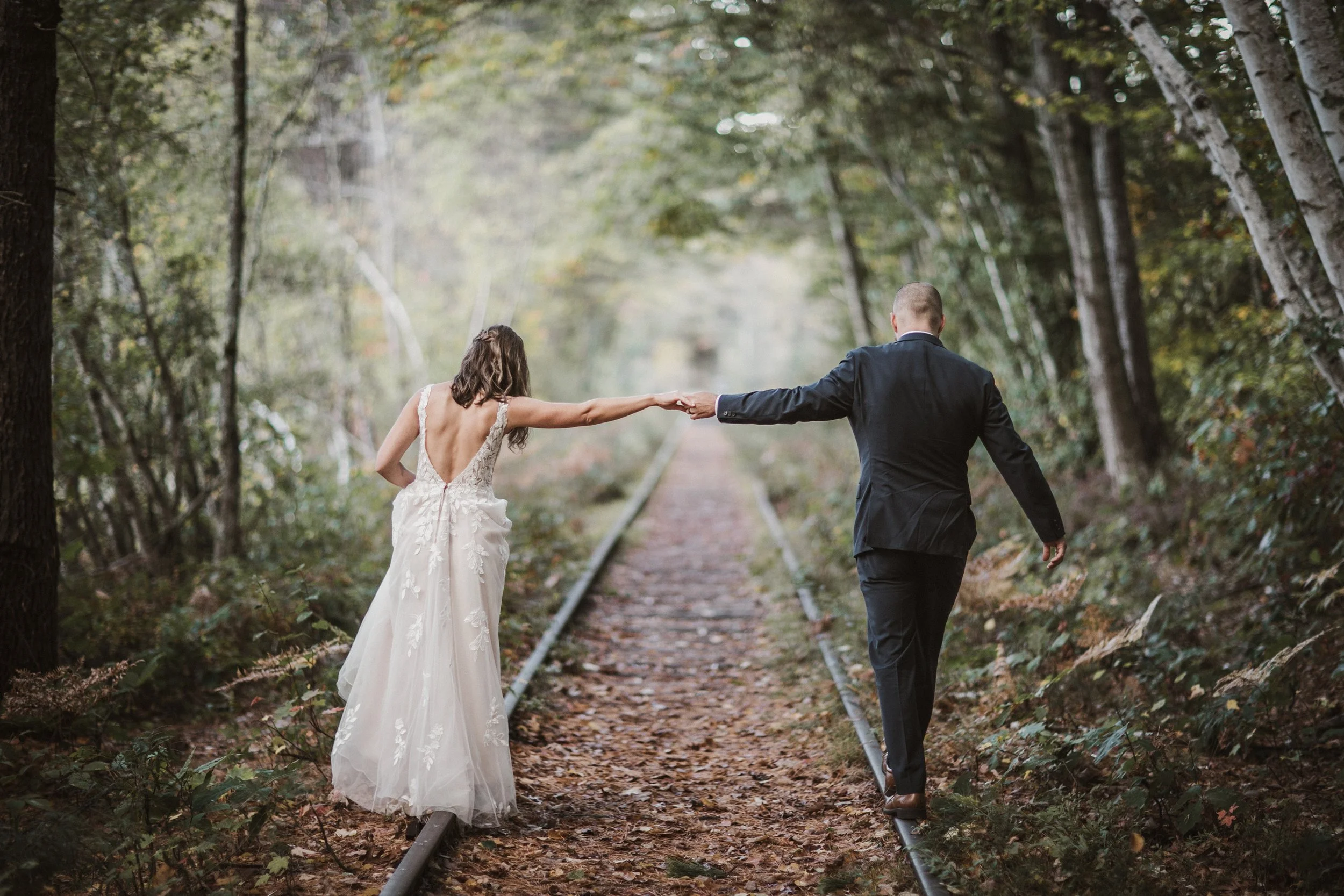 A bride and groom walking hand in hand along old railroad tracks through a forested area, surrounded by trees and foliage.