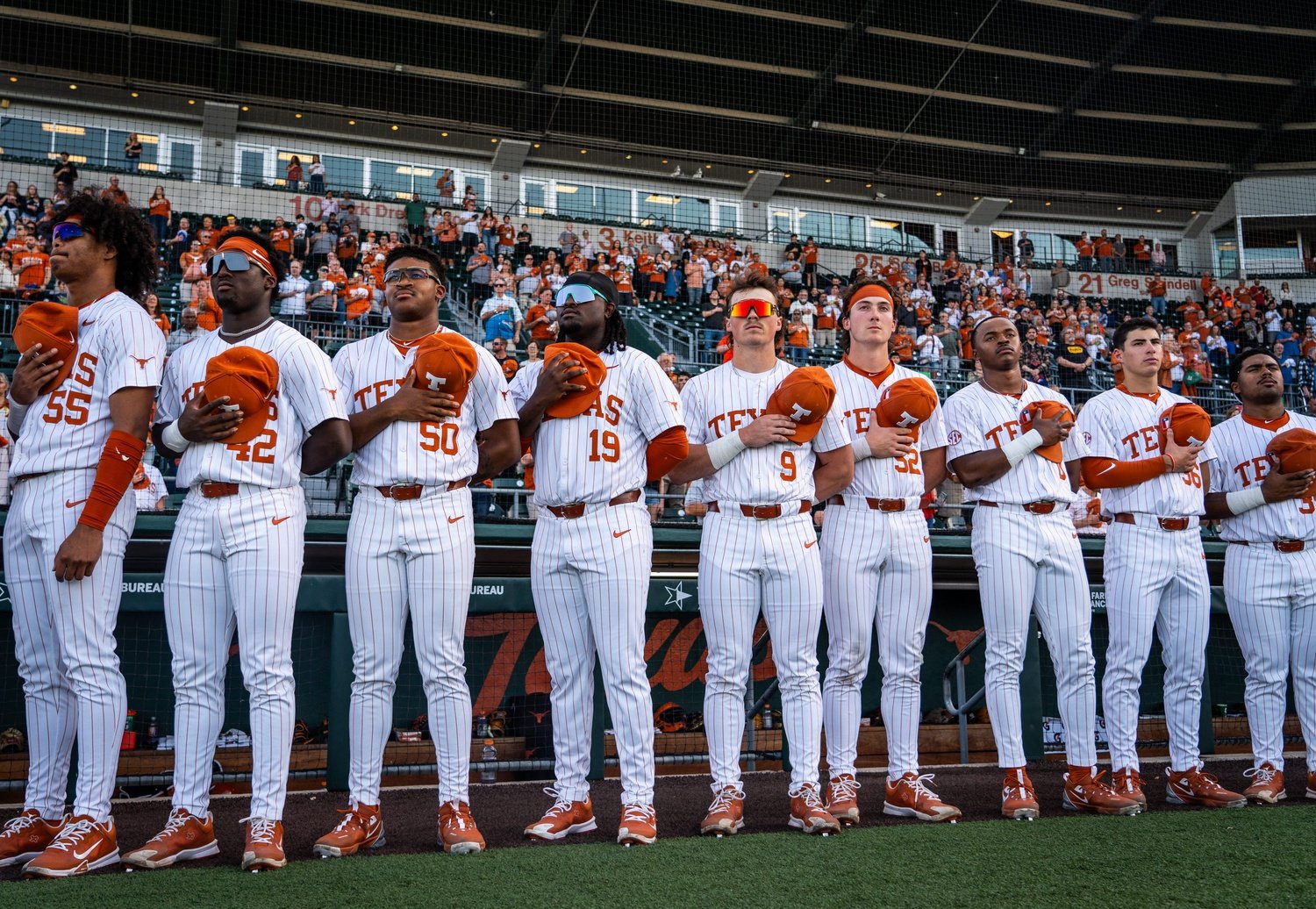 College baseball team standing with their hands over their hearts during the national anthem at a game, wearing white pinstriped uniforms with orange accents, in front of a crowd in a stadium.