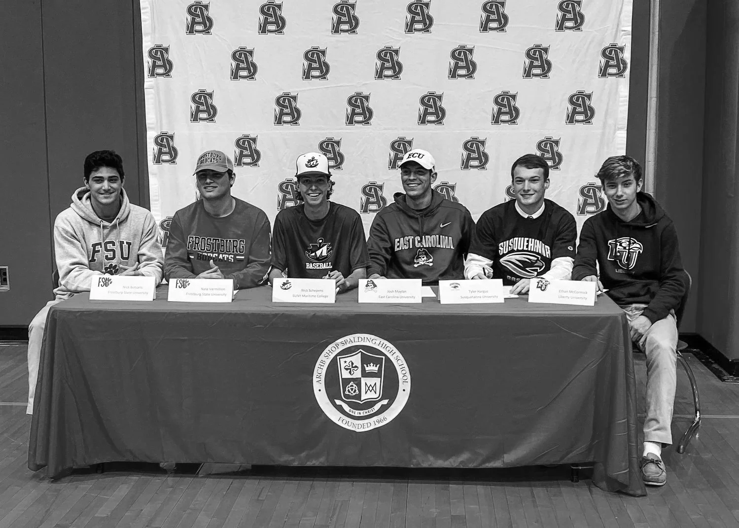 Six young men sitting at a table with nameplates, signing letters of intent for college sports, in front of a branded background and a tablecloth with a school emblem.