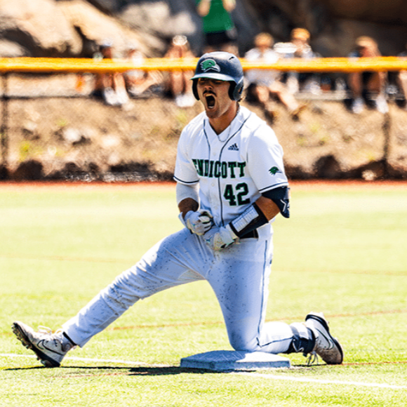 A baseball player in a white uniform with green and black accents is kneeling on the base, celebrating with his mouth open. He is wearing a helmet and batting gloves. The background shows spectators and a rocky hillside.