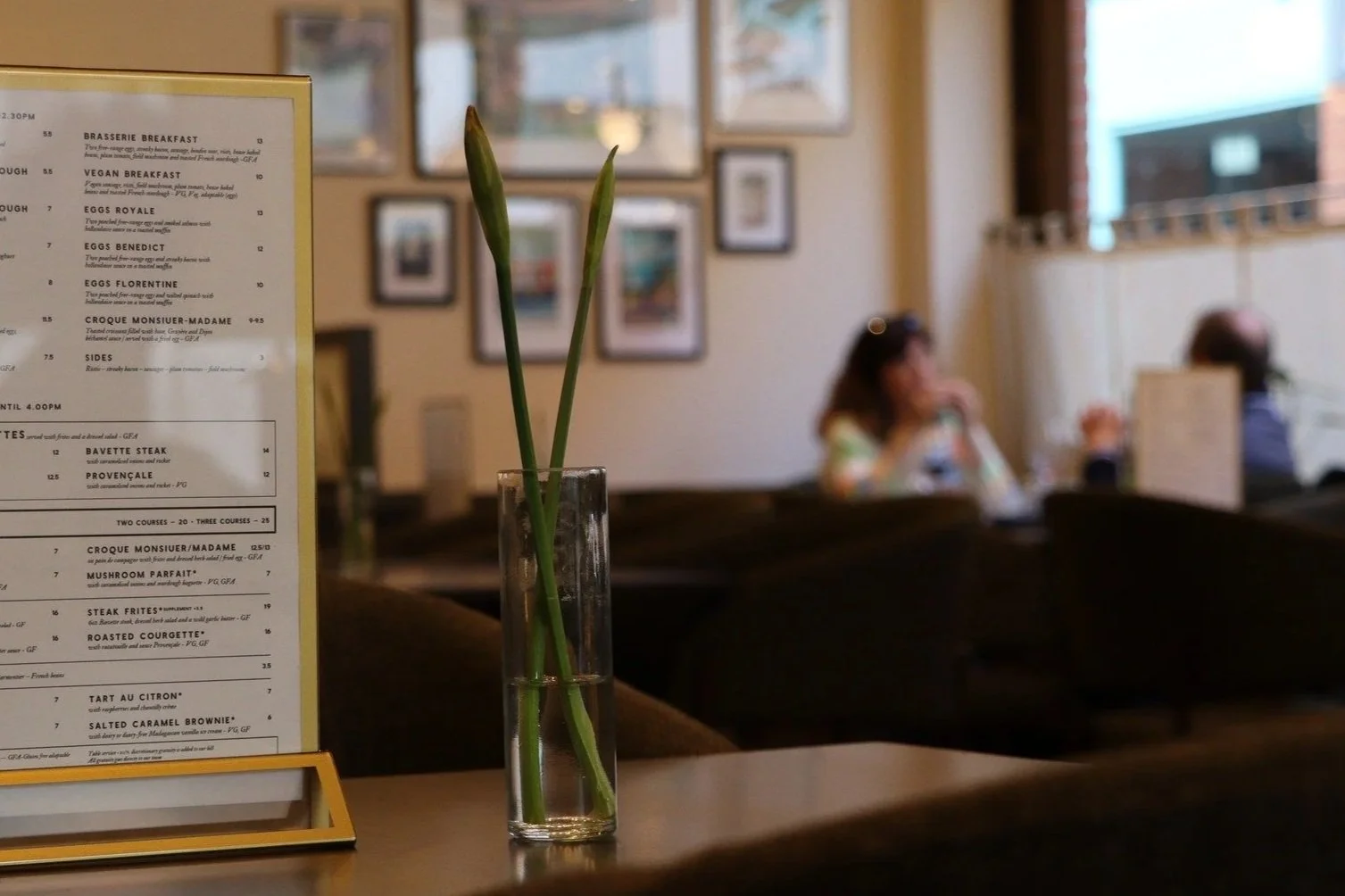A clear glass vase with water holding a single green plant stem and a closed flower bud on a table in a restaurant. A menu stand with a yellow border is visible to the left, and in the background, blurred patrons are sitting and talking in a cozy restaurant interior with framed pictures on the wall and large windows.