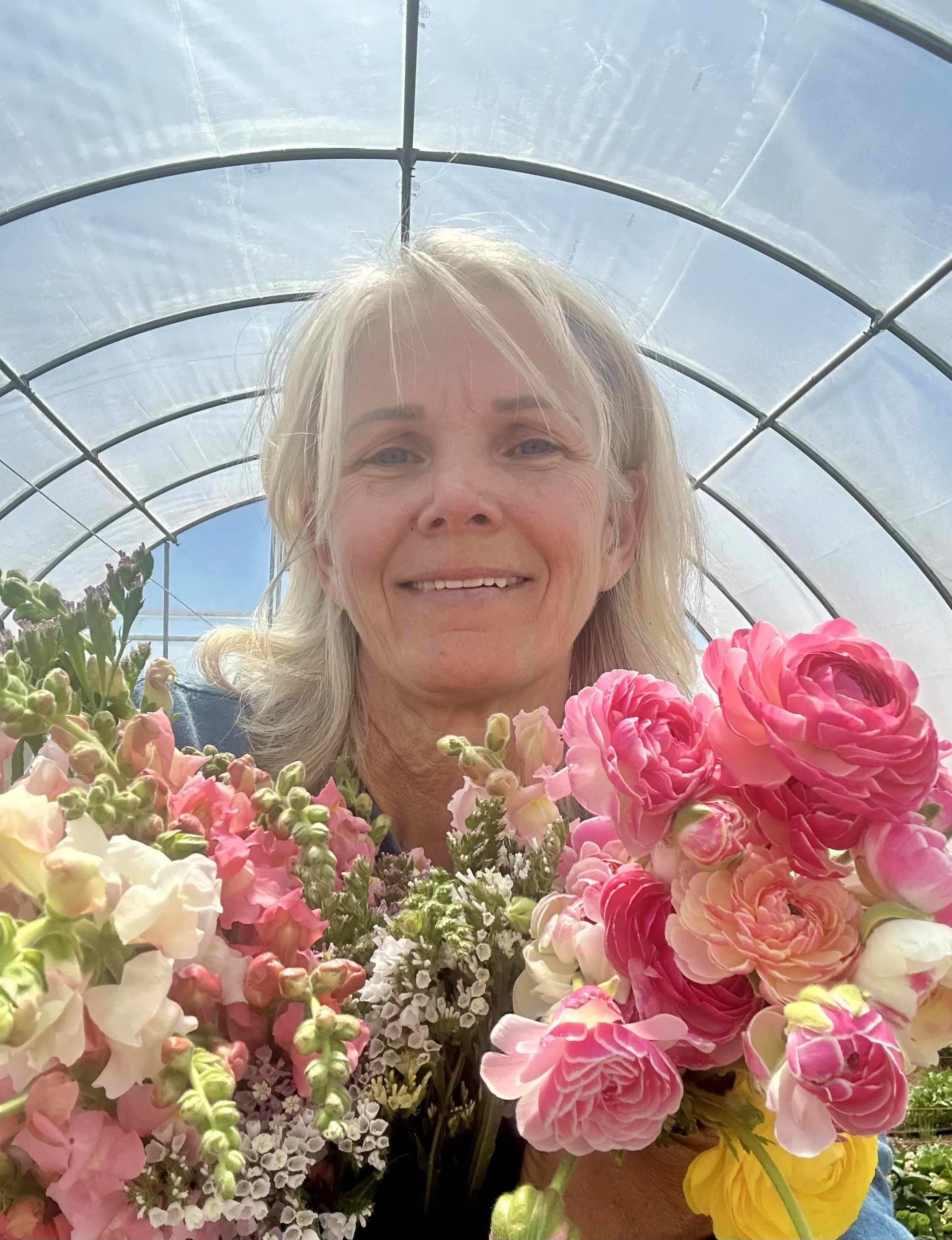 A smiling woman with shoulder-length blonde hair in a greenhouse holding a bouquet of pink, white, and yellow flowers.