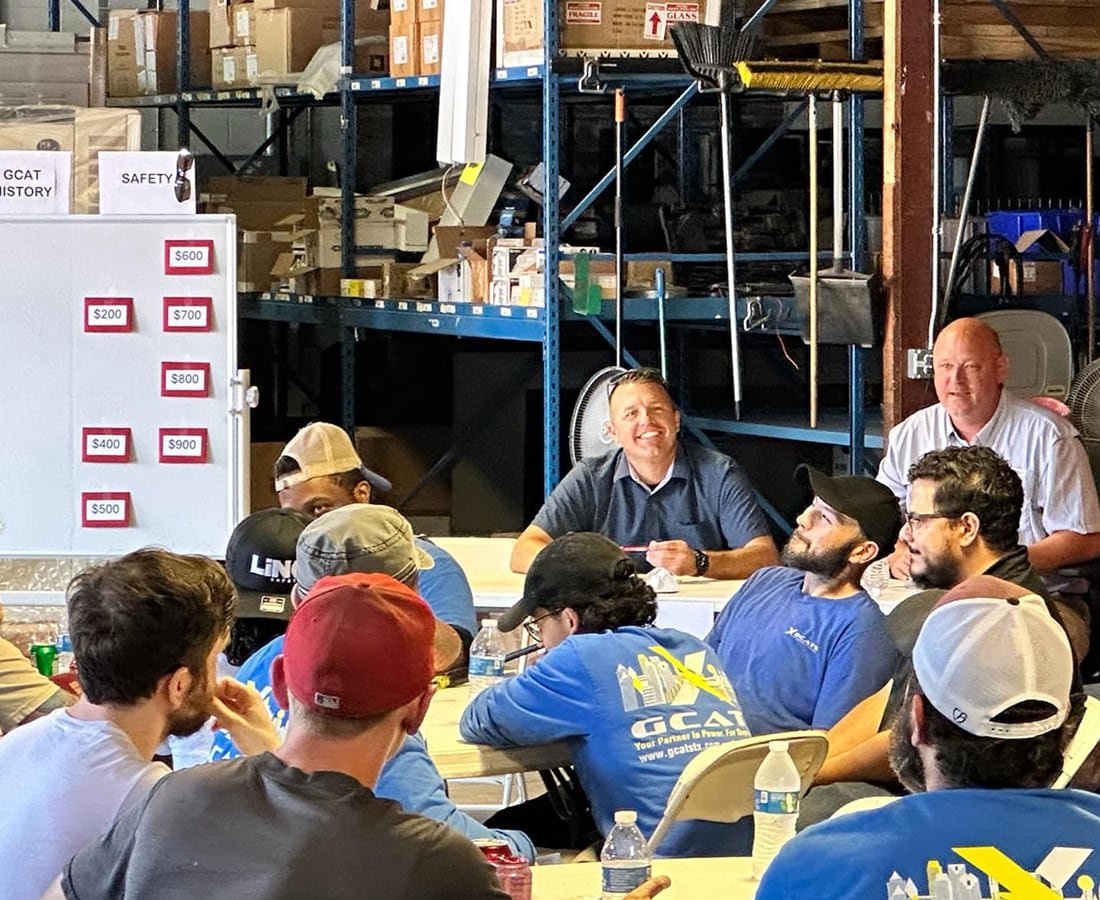 Group of people seated around a table in a warehouse, listening to two men speaking. The warehouse shelves are in the background, filled with boxes and equipment.