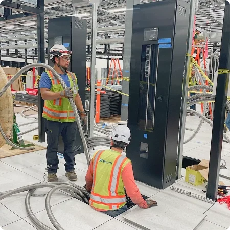 Two workers in safety vests and helmets installing or maintaining electrical equipment in an industrial facility.
