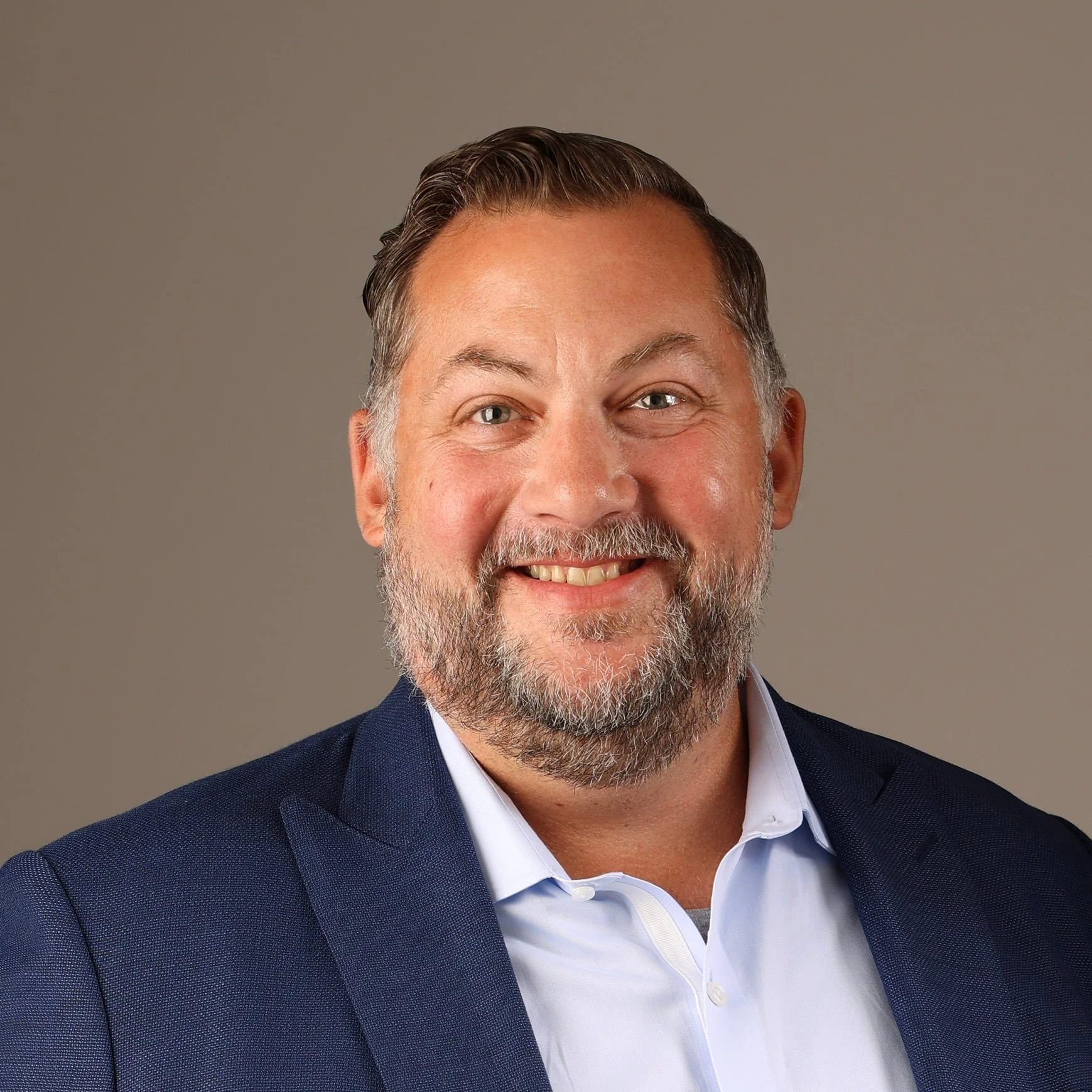 Close-up of a smiling middle-aged man with short, styled hair and a beard, wearing a navy blue suit jacket and a white collared shirt against a plain background.