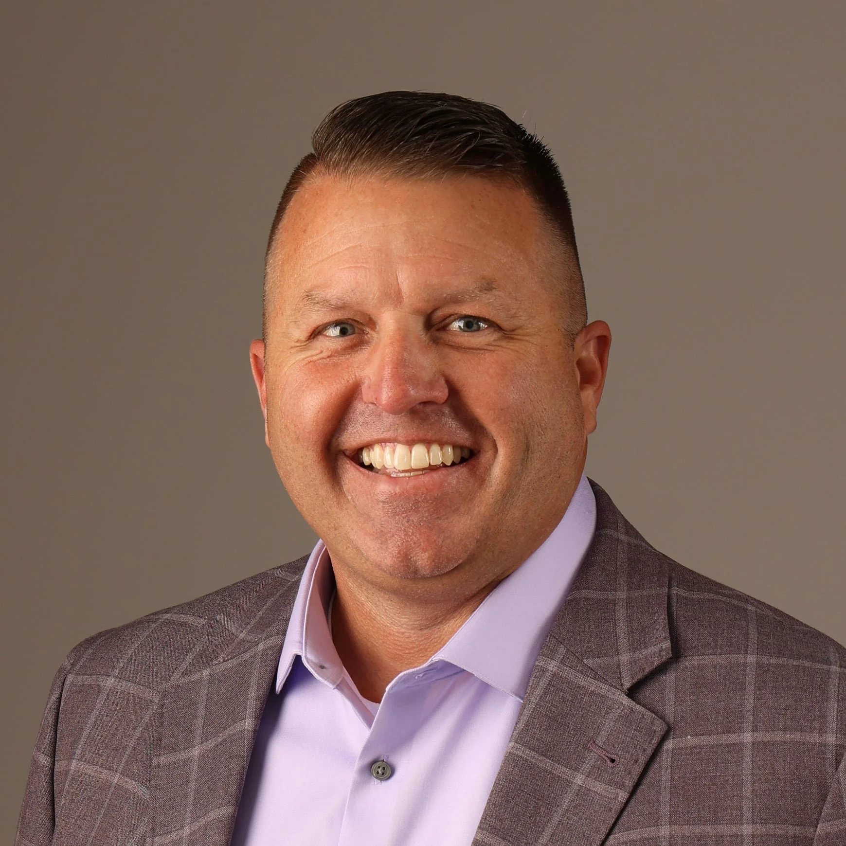 A smiling man dressed in a brown plaid suit jacket and a lavender dress shirt, with short, dark hair, posing in front of a plain gray background.