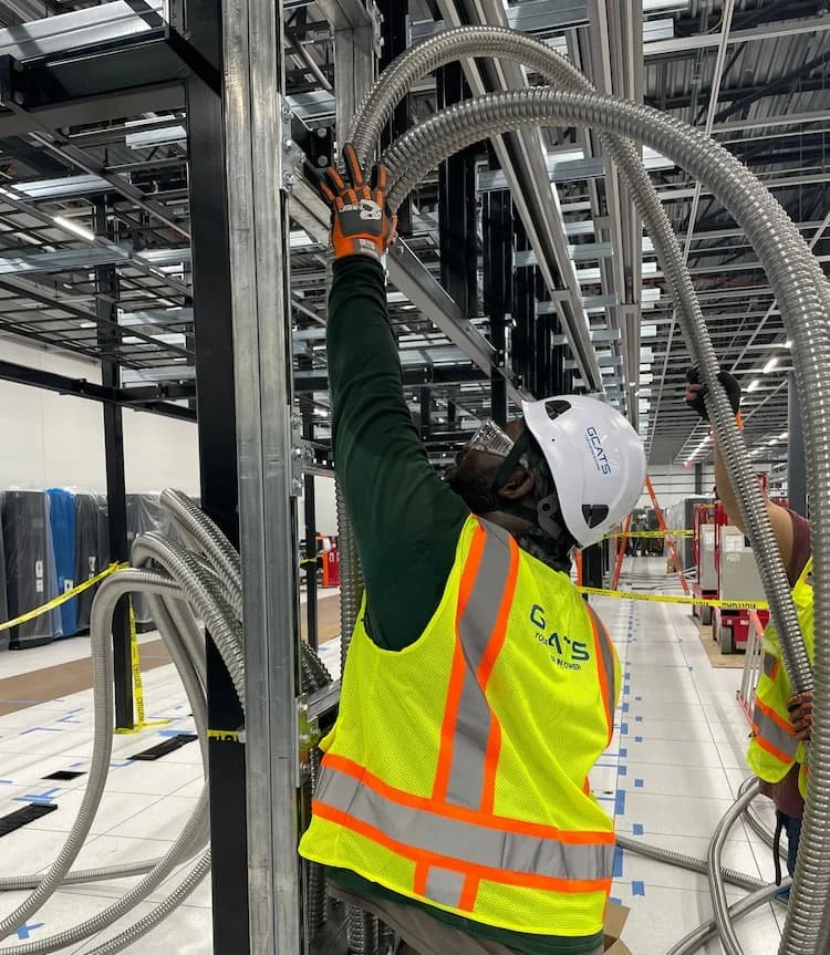 A worker in safety gear and a helmet installing or repairing electrical wiring or cabling in a large industrial or data center environment.