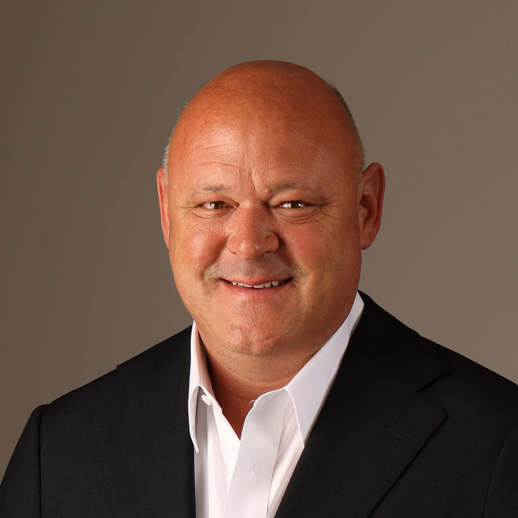 Close-up of a smiling bald man wearing a black suit and white shirt against a plain background.