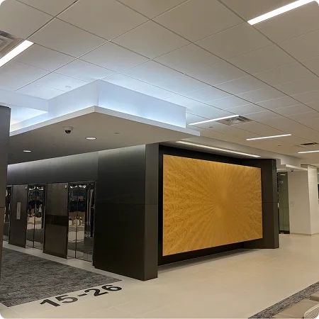 Modern indoor space with a large digital display attached to a black wall, featuring a wooden pattern background. The ceiling has recessed lighting, and there are glass doors or windows on the left side. The floor is light-colored with a section of carpet near the display.