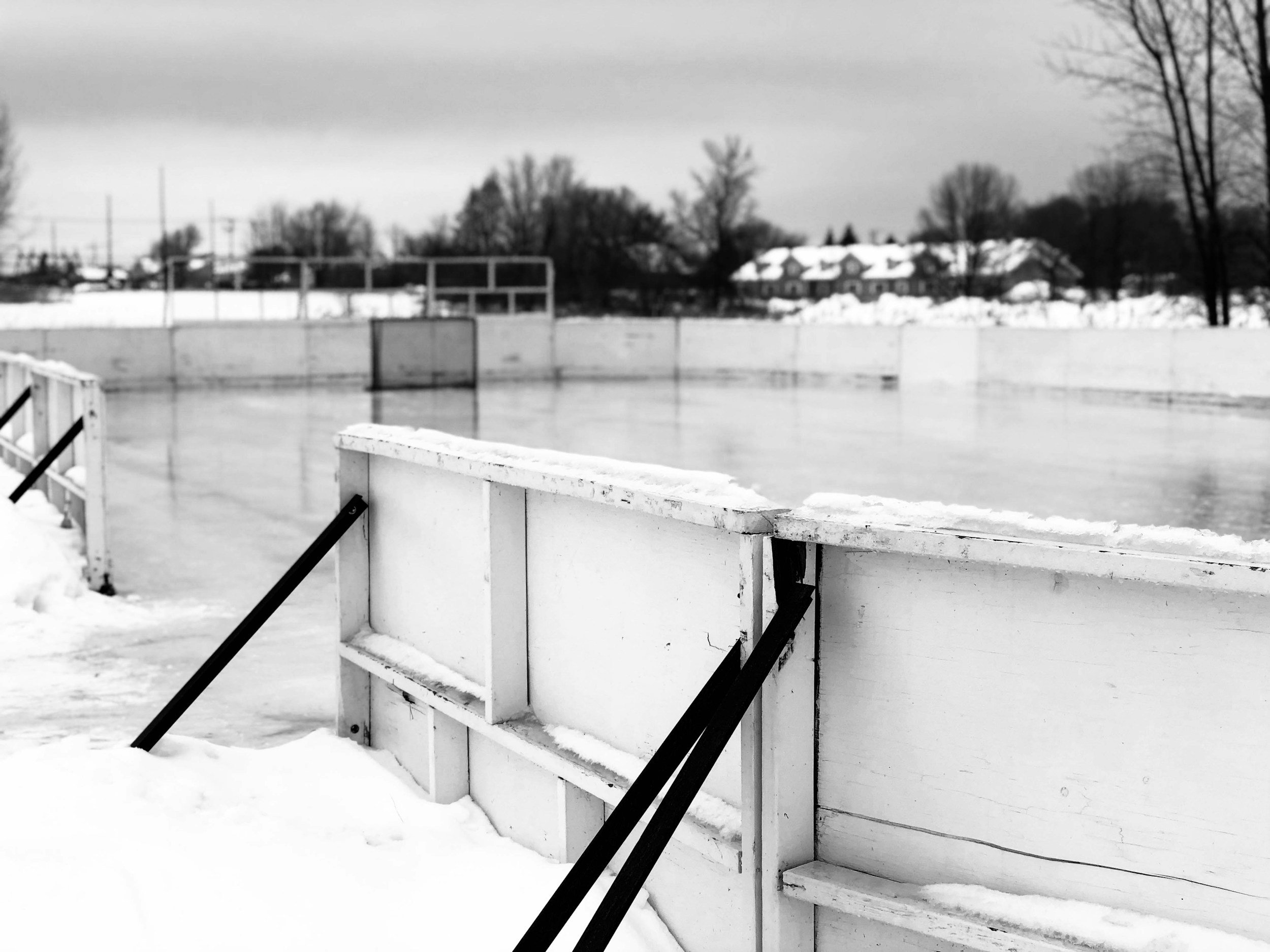An outdoor ice hockey rink covered in snow with a rink barrier in the foreground, against a wintery cloudy sky and snow-covered trees and houses in the background.