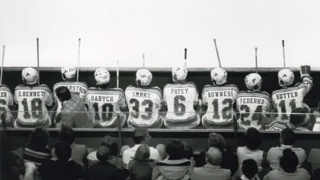 Hockey players sitting on a bench with helmets and jerseys, fans seated below watching the game.