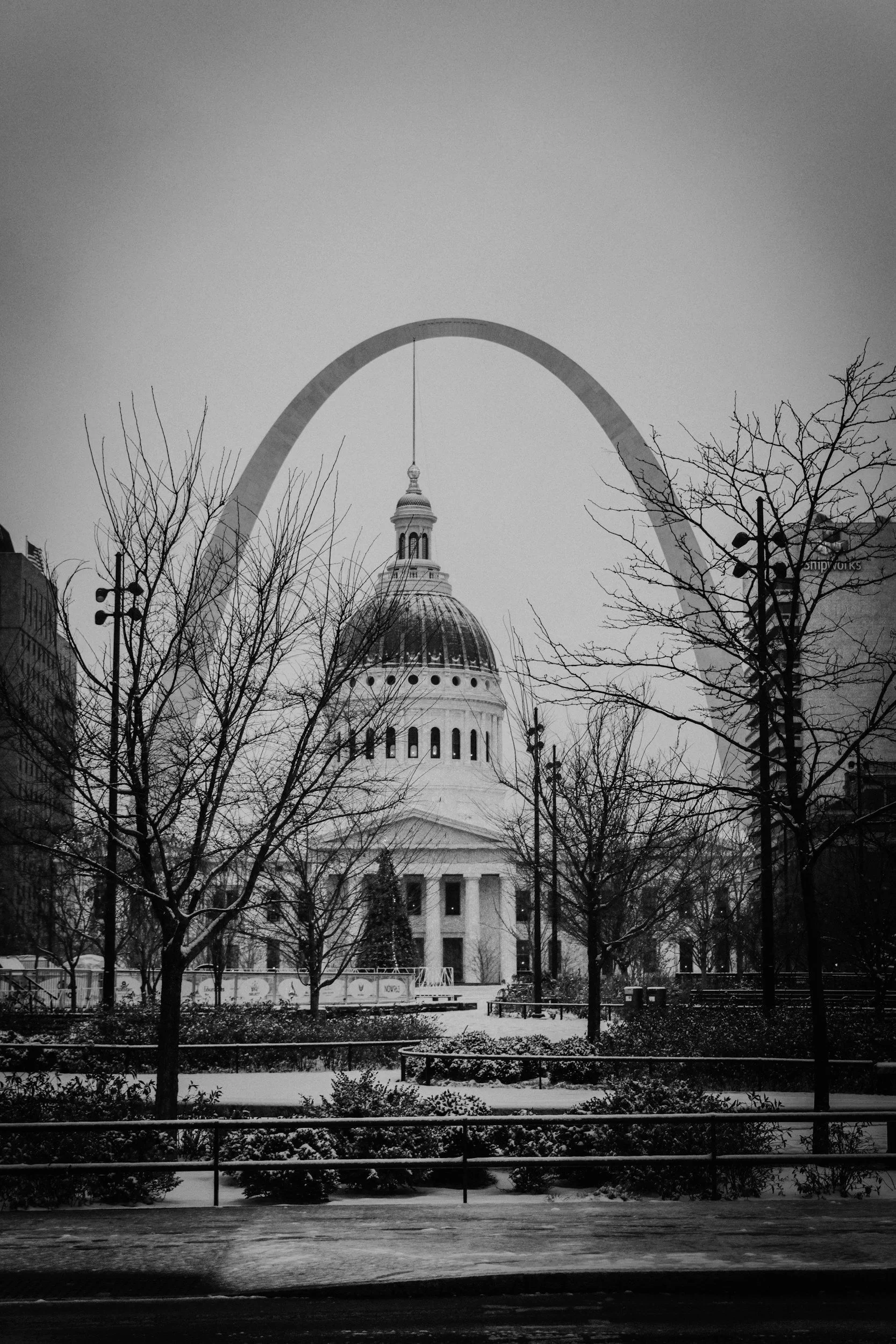 Black and white photo of a city park with trees and bushes covered in snow, a large dome building in the background, and the Gateway Arch in St. Louis visible behind the building.